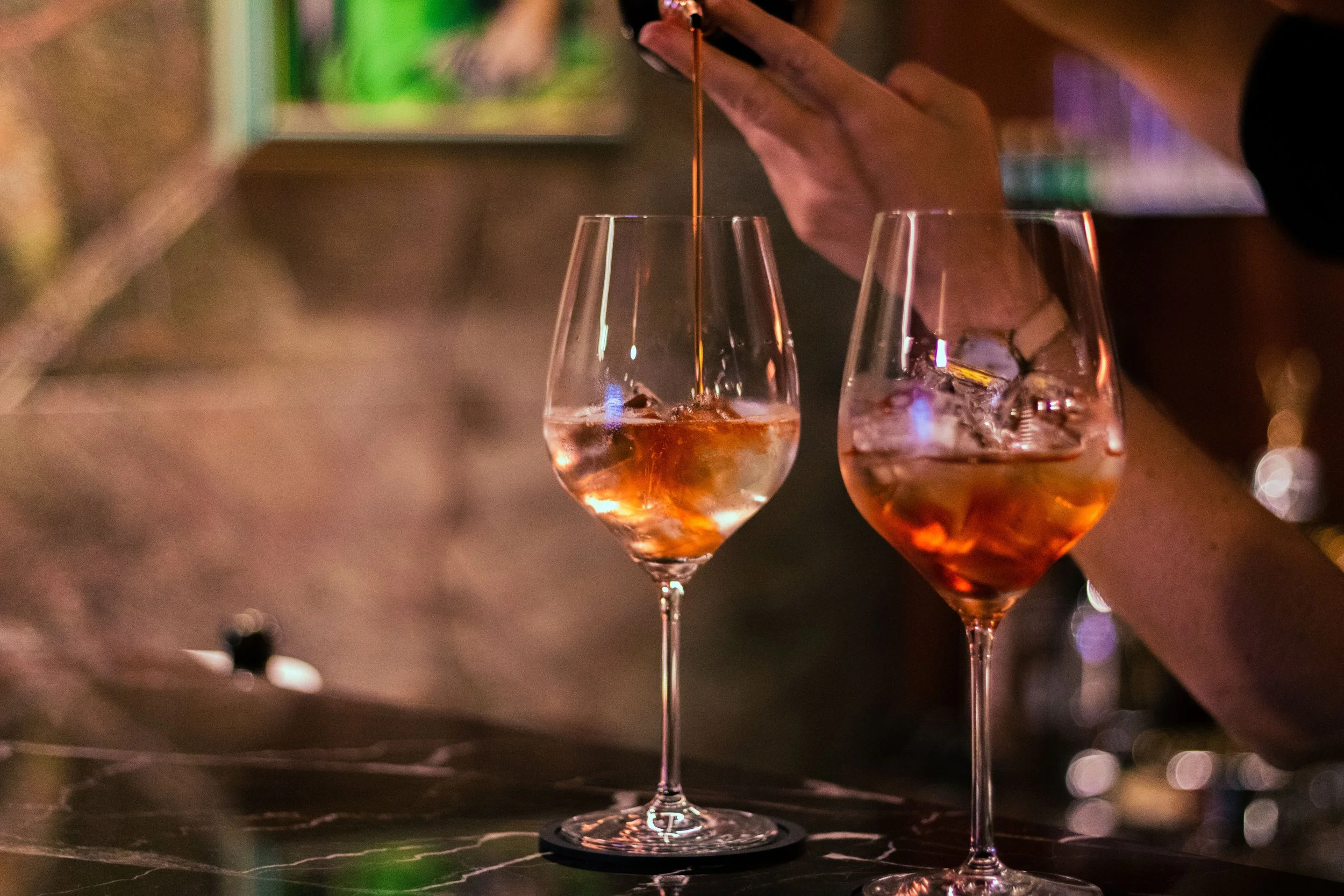 A bartender pouring a drink into a wine glass with ice cubes, while another glass with ice sits on the bar counter. The scene is dimly lit, with a blurred background.