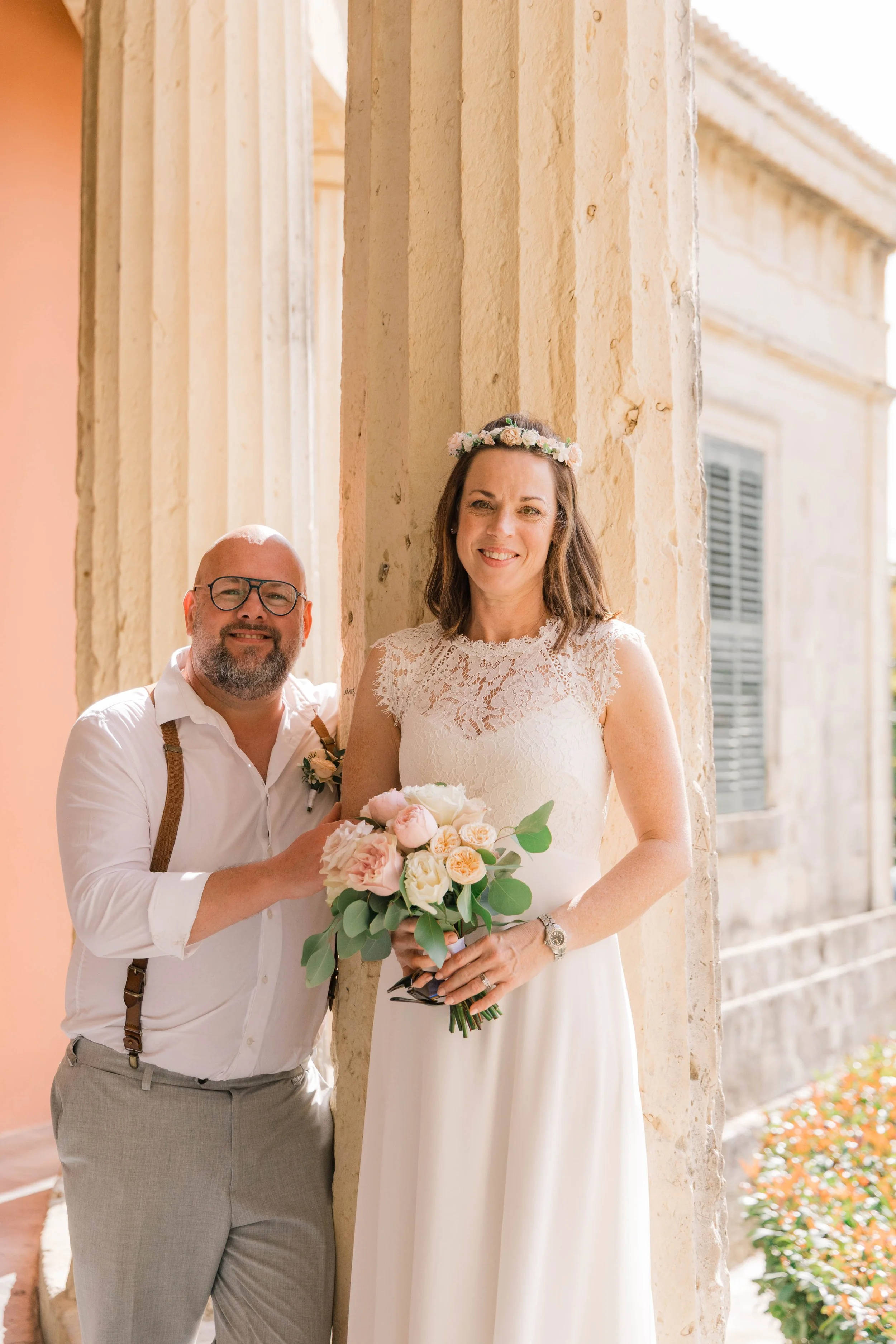 A smiling woman in a white wedding dress with a flower crown holding a bouquet of pink and white flowers, standing next to a man in a white shirt and glasses, outside beside a beige stone wall.