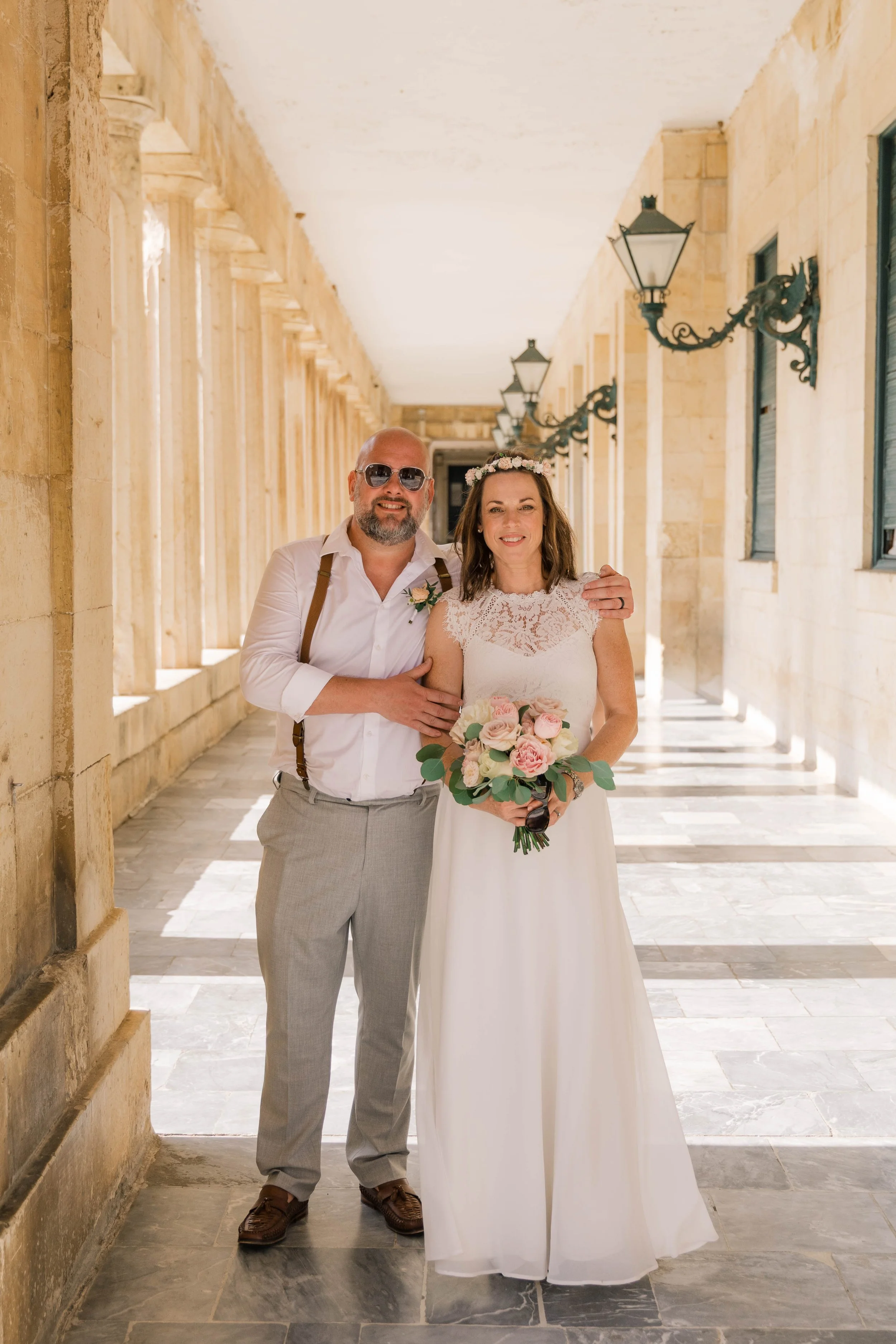 A woman in a white wedding dress holding a bouquet of pink and white roses and a man in a white shirt and gray pants standing together in a sunlit stone corridor, with the man placing his arm around the woman's shoulders.