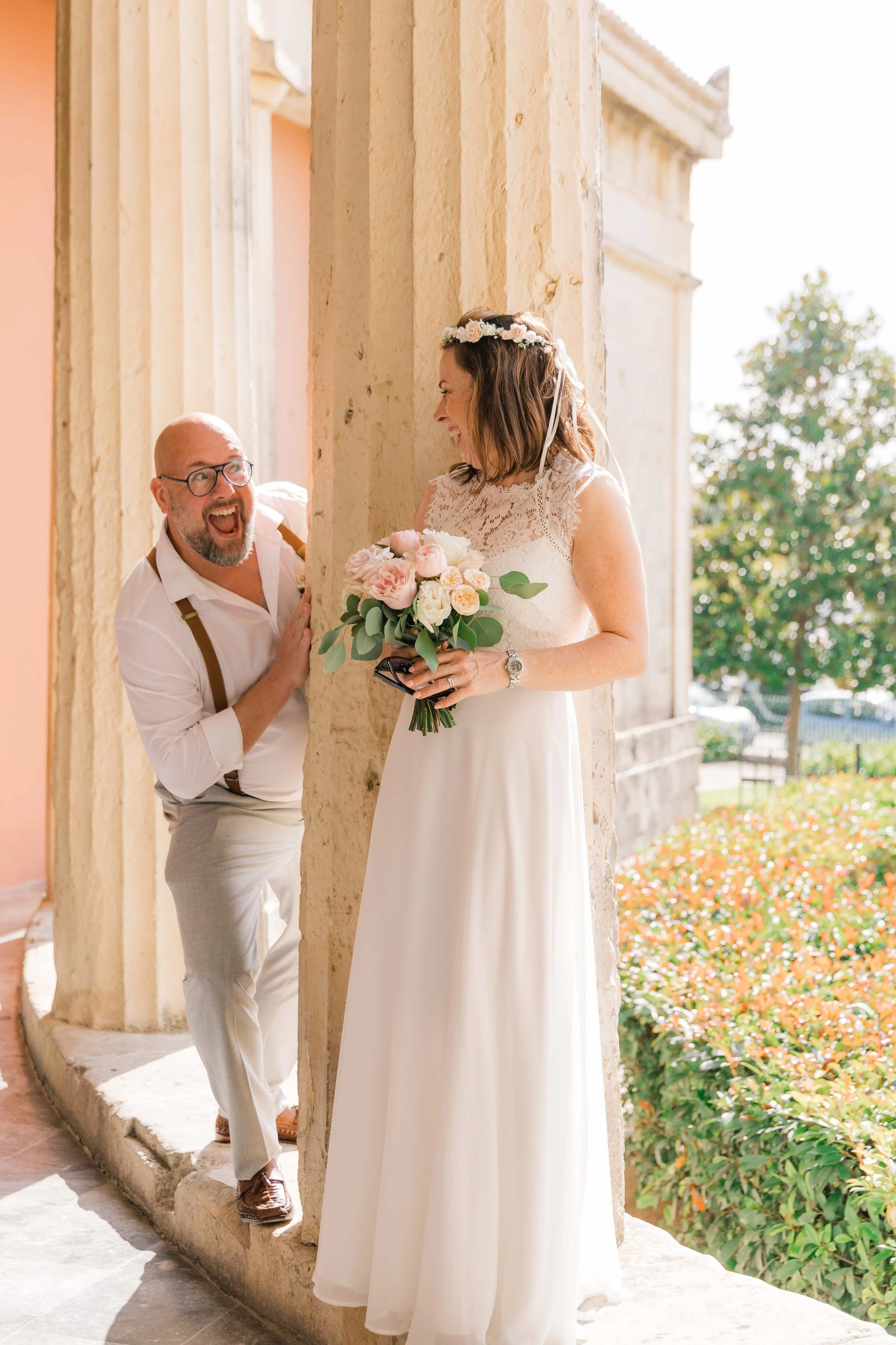A woman in a wedding dress holding a bouquet laughing as a man peeks from behind a stone pillar outside a building.