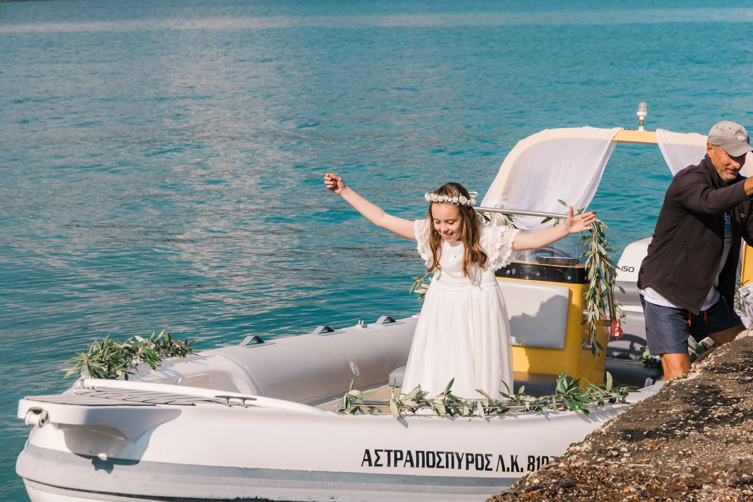 A young girl wearing a white dress and floral headband standing on a decorated boat near the water edge with arms raised in celebration.