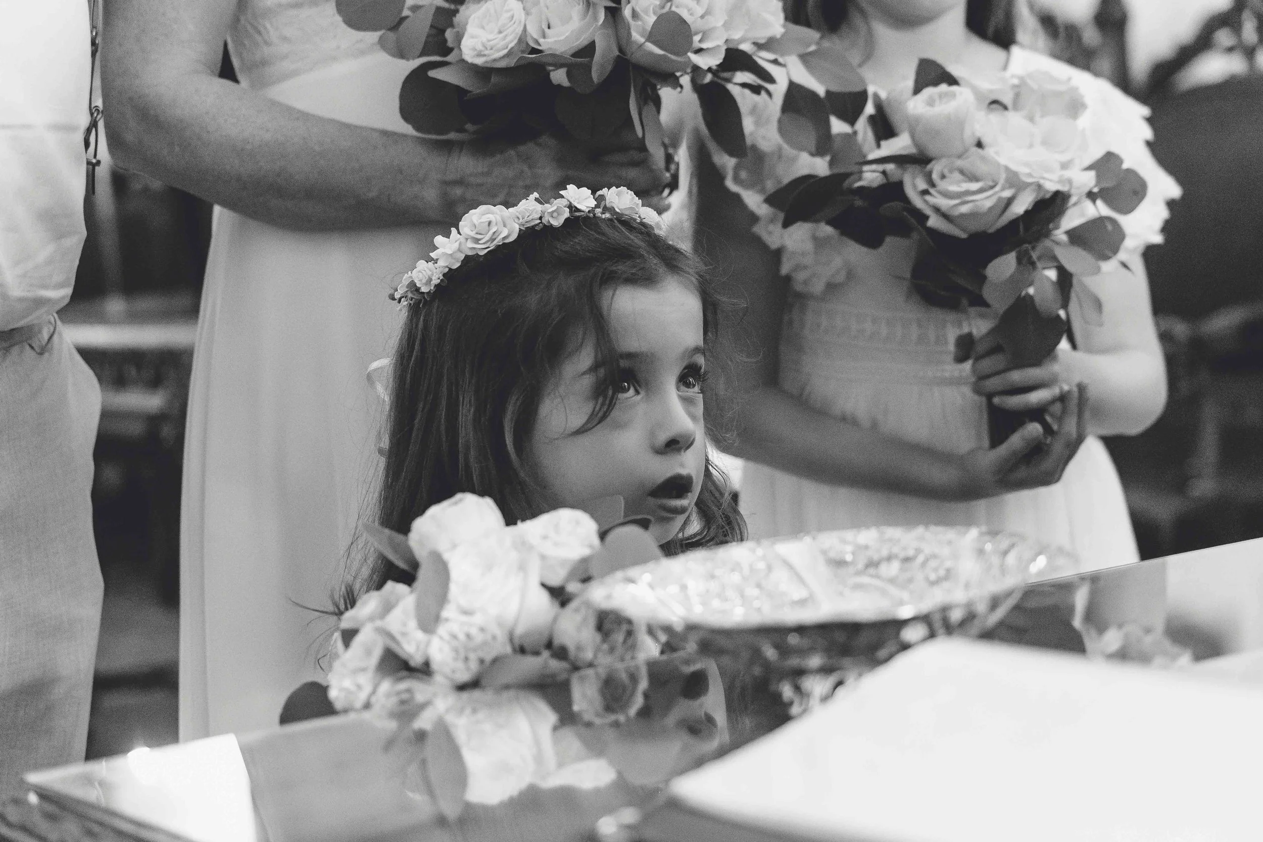 A young girl with a floral crown looks up with a surprised or curious expression, surrounded by bouquets of flowers at what appears to be a celebration or special event.