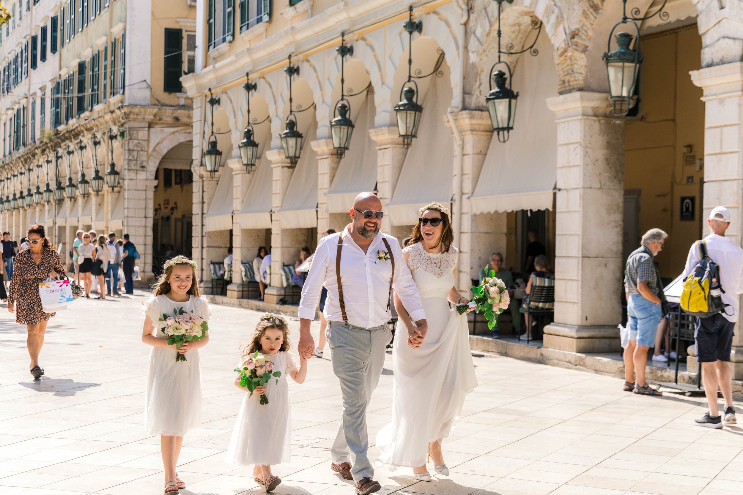 A wedding procession walking along a sunlit European city street with historic buildings. The bride is in a white dress holding a bouquet and is walking hand-in-hand with the groom, who is dressed in a white shirt with suspenders. Two young girls in 