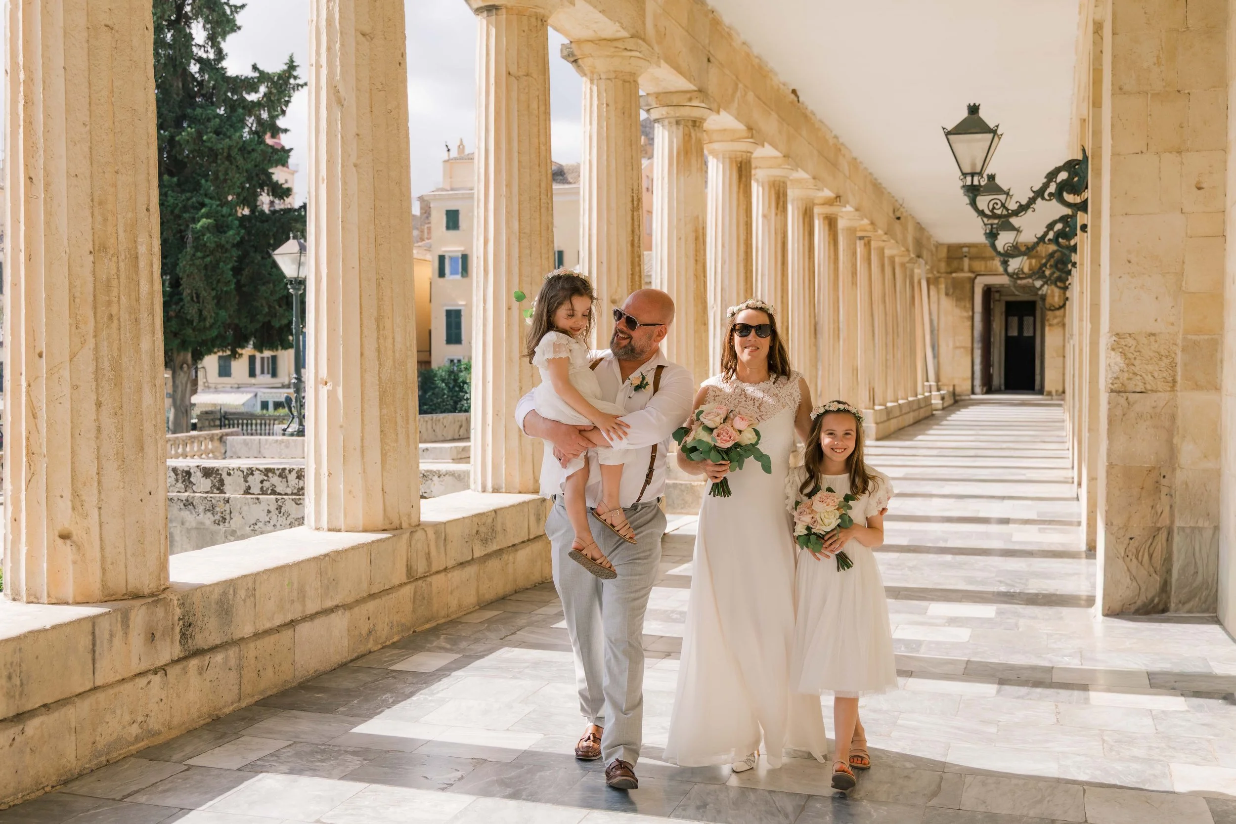 A wedding party walking through a colonnade. The bride is holding a bouquet, accompanied by a man and two young girls holding smaller bouquets. The group is smiling and dressed in white, with the setting featuring large stone columns and decorative l