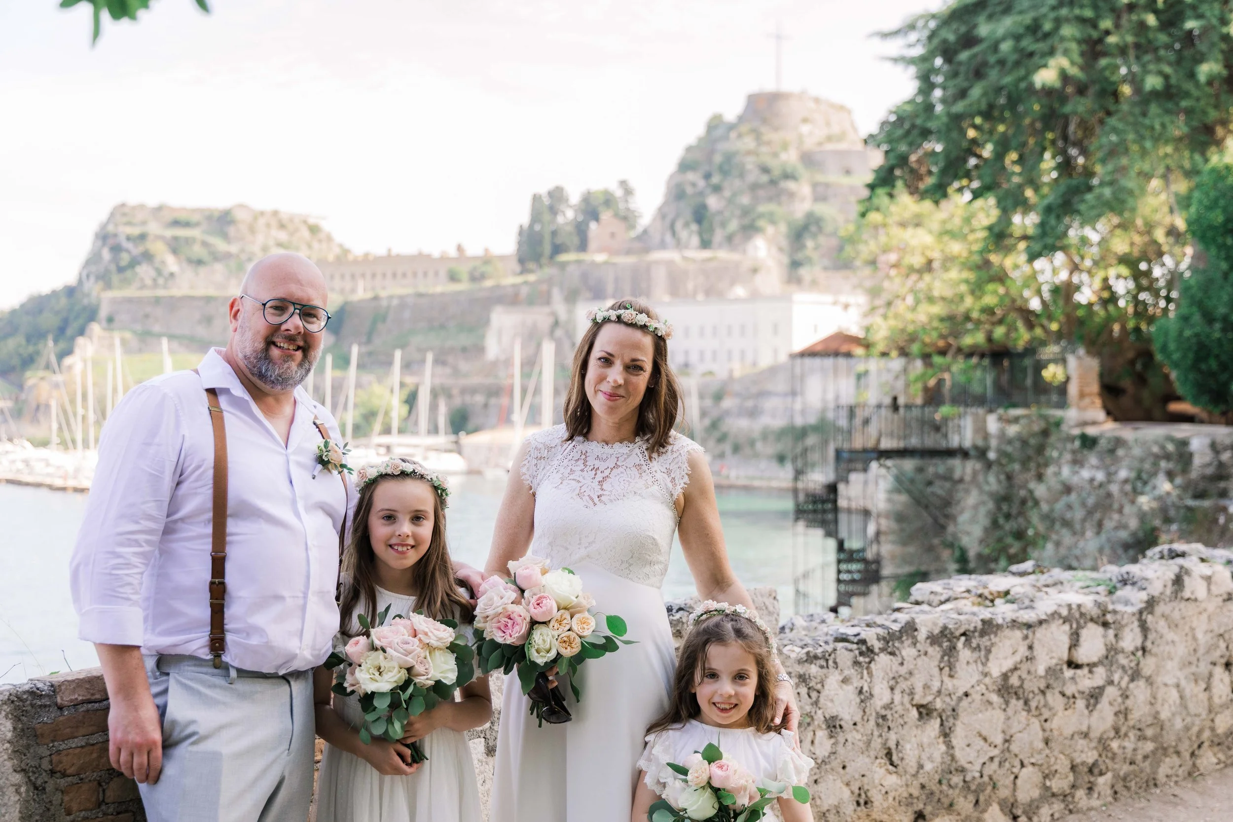 A family of four celebrating at an outdoor wedding near a body of water with boats. The man is wearing glasses, a white shirt, and suspenders. The woman and two girls are dressed in white, with floral crowns and holding bouquets of pink and white ros