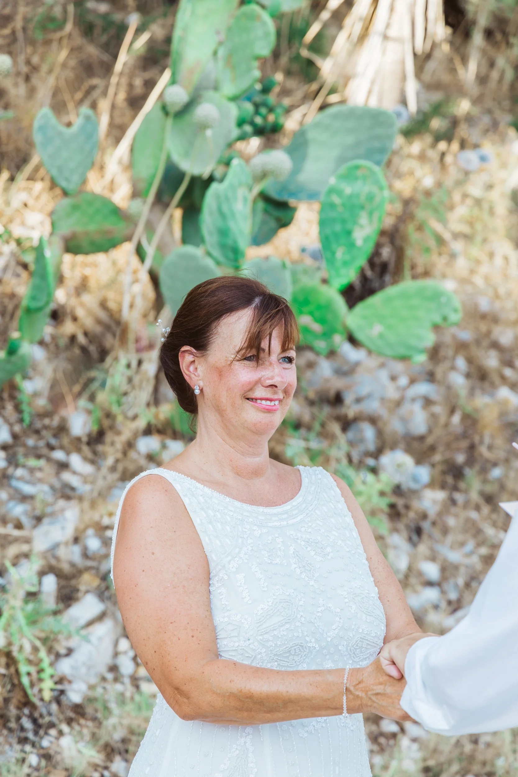 A woman in a white wedding dress with short brown hair, smiling and shaking hands with someone outside, in front of a cactus plant.