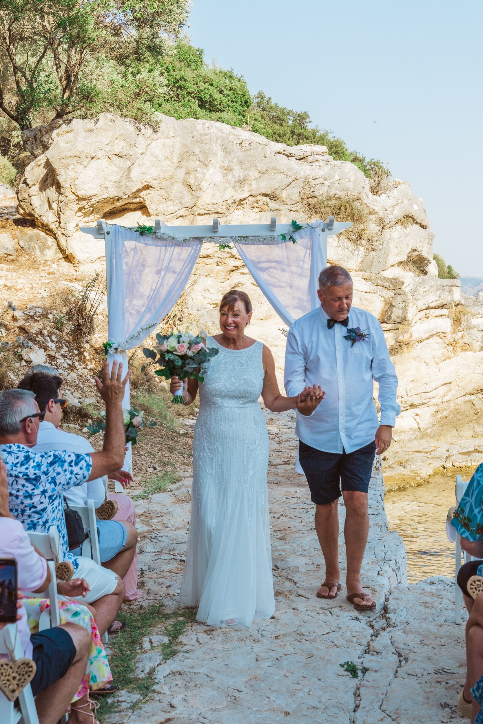 Couple walking down the aisle after getting married at a beachside wedding ceremony, with an audience seated on either side, and a rocky cliff background under a white canopy with sheer drapes and greenery.