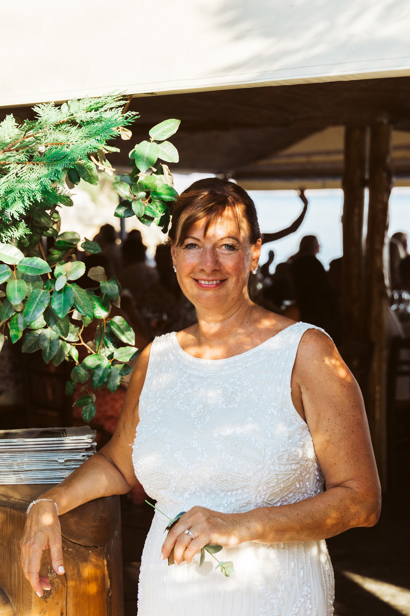 Woman in white dress smiling at a celebration in a decorated outdoor venue with guests in the background.