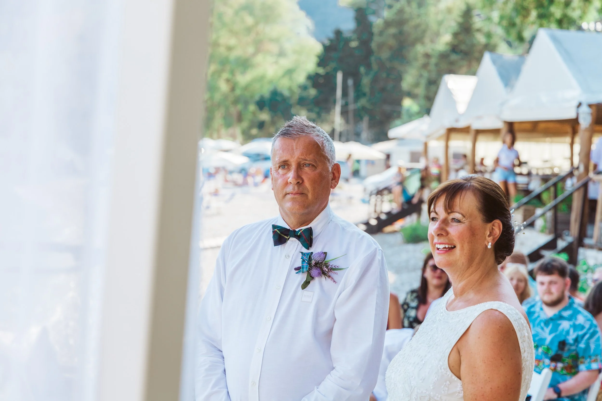 A bride and groom stand during their outdoor wedding ceremony, with guests seated in the background and a white tent in the background.