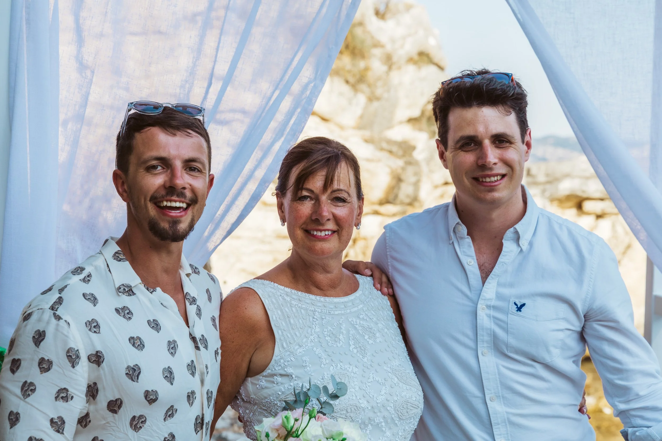 Three people, two men and one woman, smiling outdoors near a blue fabric backdrop with a rocky landscape in the background. The woman holds a small bouquet of flowers.