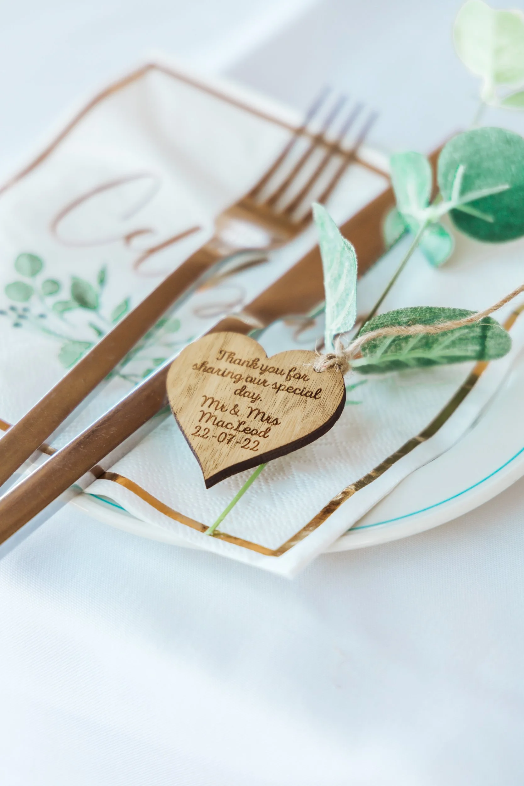 Wedding place setting with a fork, knife, a heart-shaped wooden tag reading "Thank you for sharing our special day, M & M Macleod 22-07-22", green leaves, and a white napkin with gold accents.