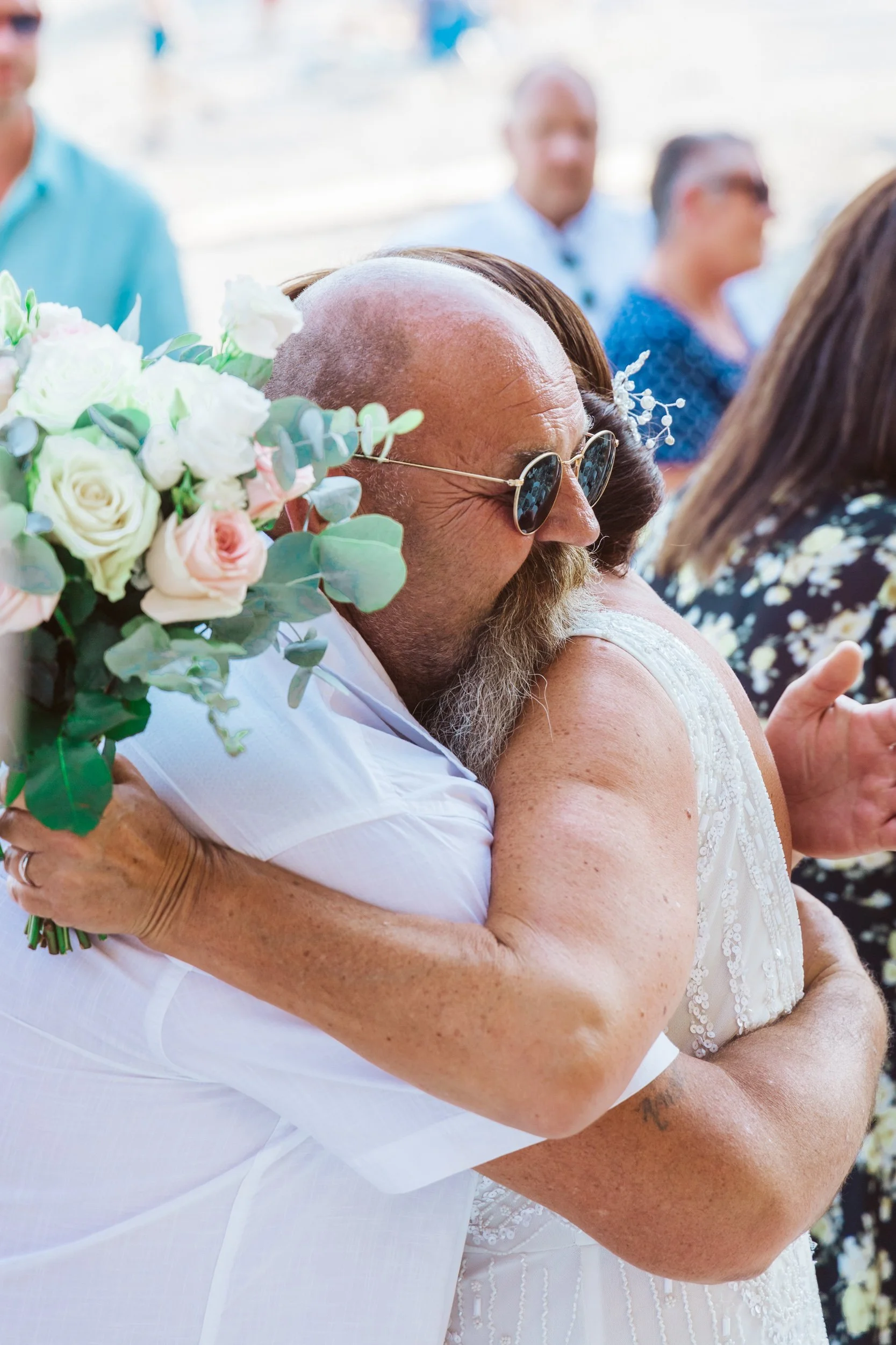 A man with sunglasses and a beard hugging a woman in a wedding dress, holding a bouquet of white and pink roses with greenery, at a wedding reception.