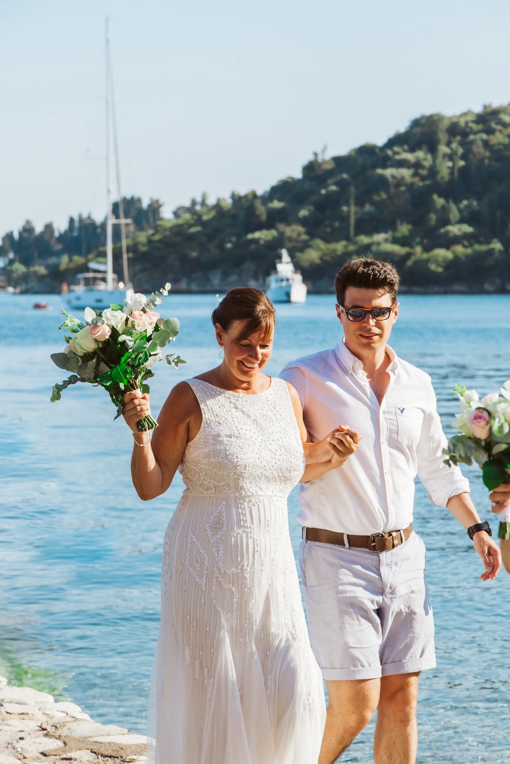 A woman in a white lace dress holding a bouquet and smiling walking alongside a man in a white shirt, shorts, and sunglasses by a water body with boats and a mountain in the background.