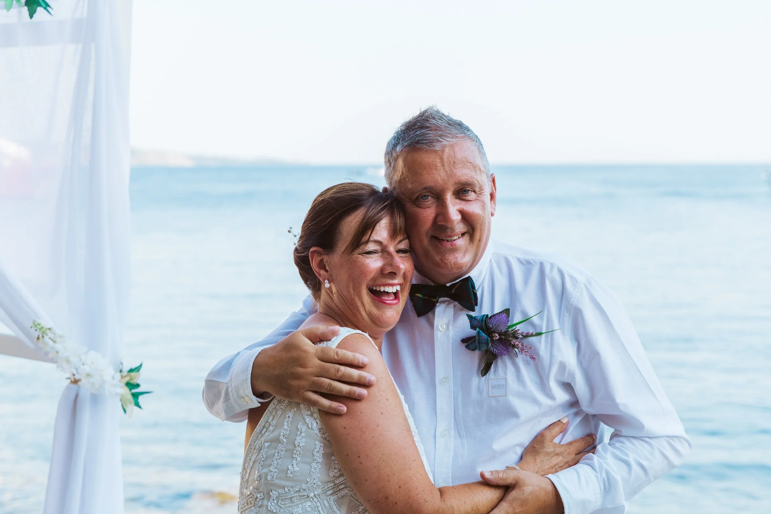 A happy couple dressed in wedding attire embracing by the water with a white beach wedding arch in the background.