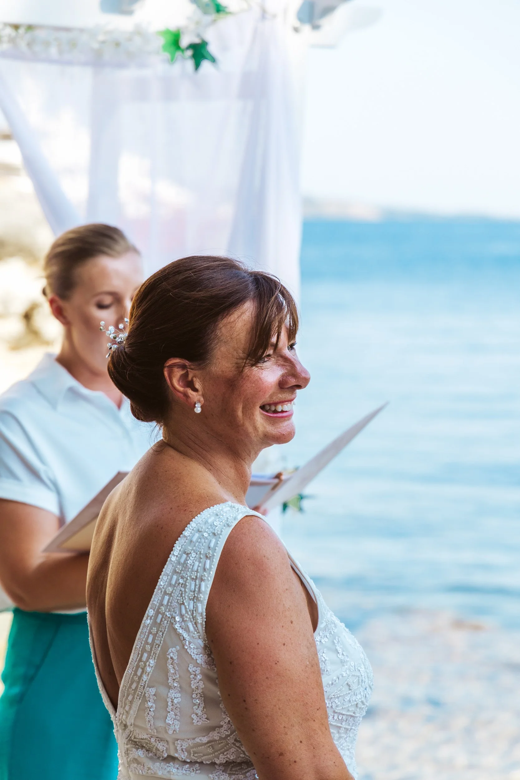 A woman in a wedding dress smiling during a beach wedding ceremony with an officiant in the background, near the water.
