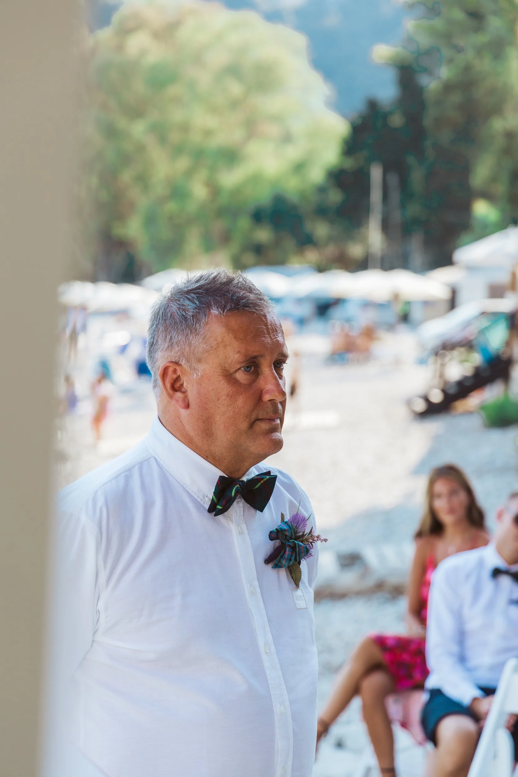 A groom standing outdoors at a beach wedding, wearing a white dress shirt, a plaid bow tie, and boutonniere, with a serious expression, blurry bride and guests in the background.