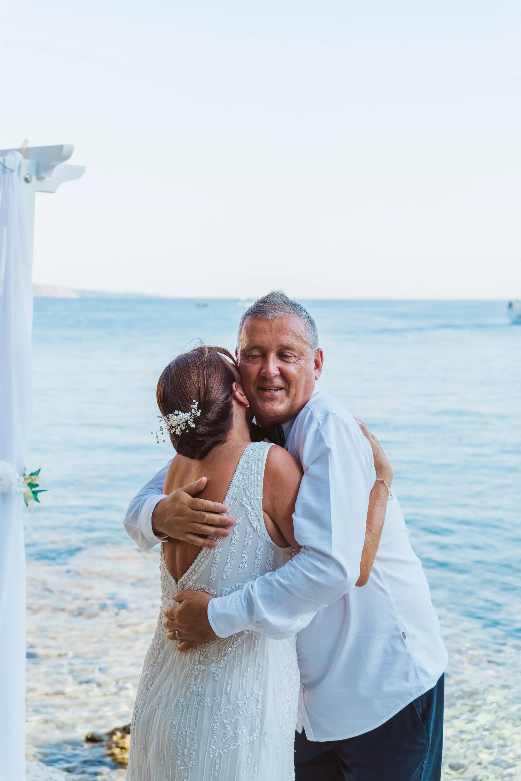 A bride and groom share a hug during a wedding ceremony by the water, with the ocean in the background and clear blue sky overhead.