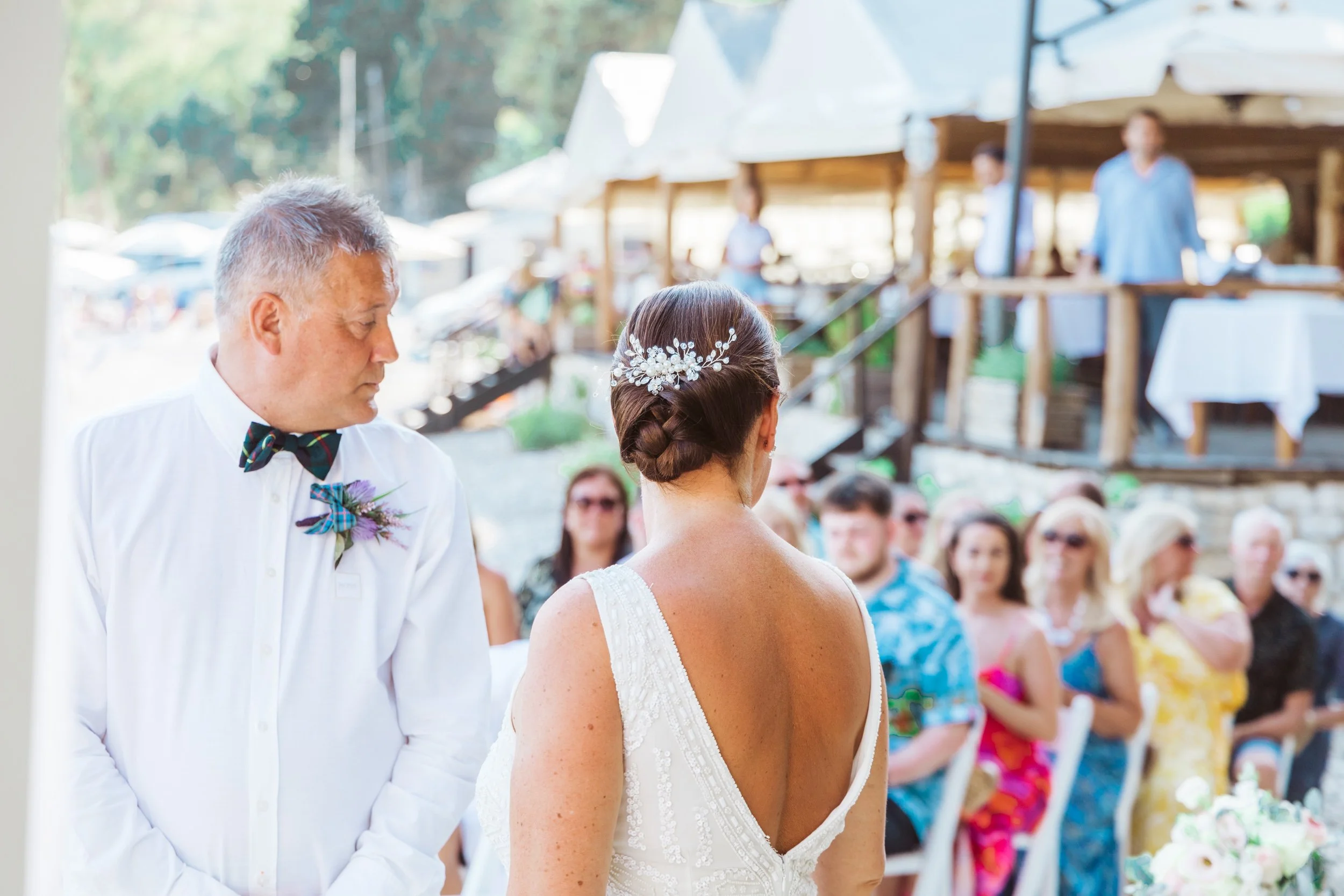 A bride and an older man in a wedding ceremony outdoors, with guests seated and standing in the background, some wearing colorful attire, at a rustic venue with wooden structures.