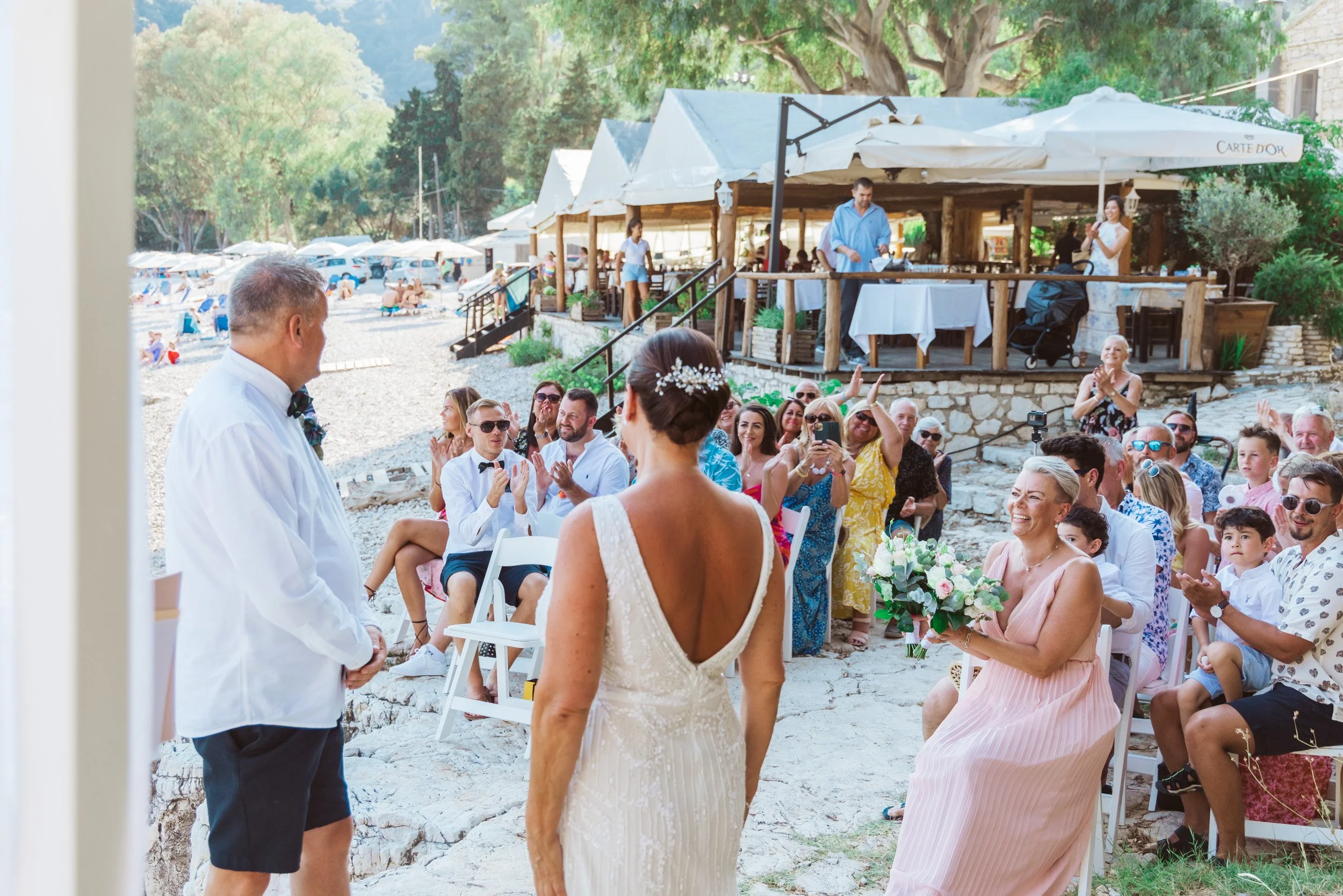 A wedding ceremony outdoors on a rocky beach with guest seated and standing, clapping and smiling, as the bride and groom face each other.