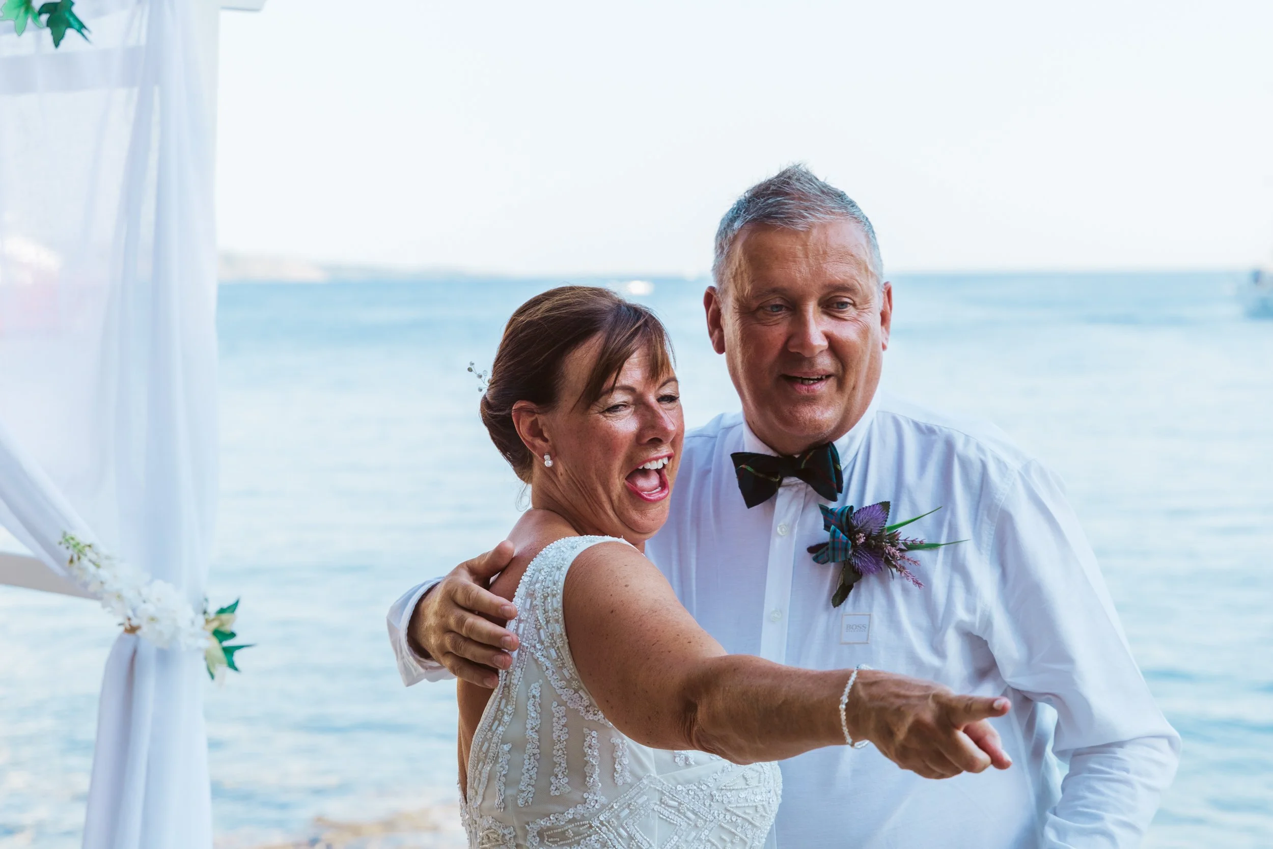 A joyful couple dancing at their wedding by the water with a white draped canopy and floral decorations.