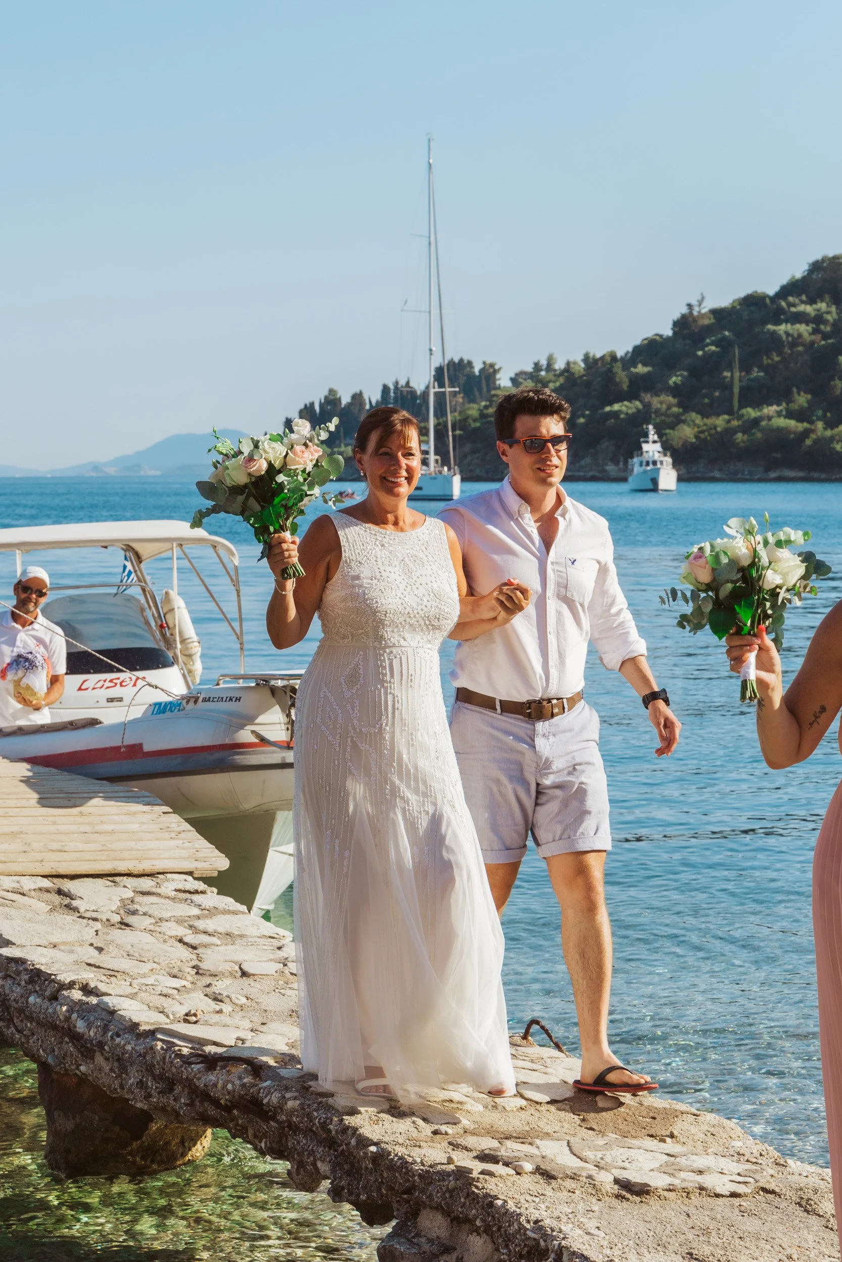A woman in a white dress and a man in a white shirt with shorts walking on a stone dock by the water, holding bouquets of flowers, with boats and a hilly shoreline in the background.