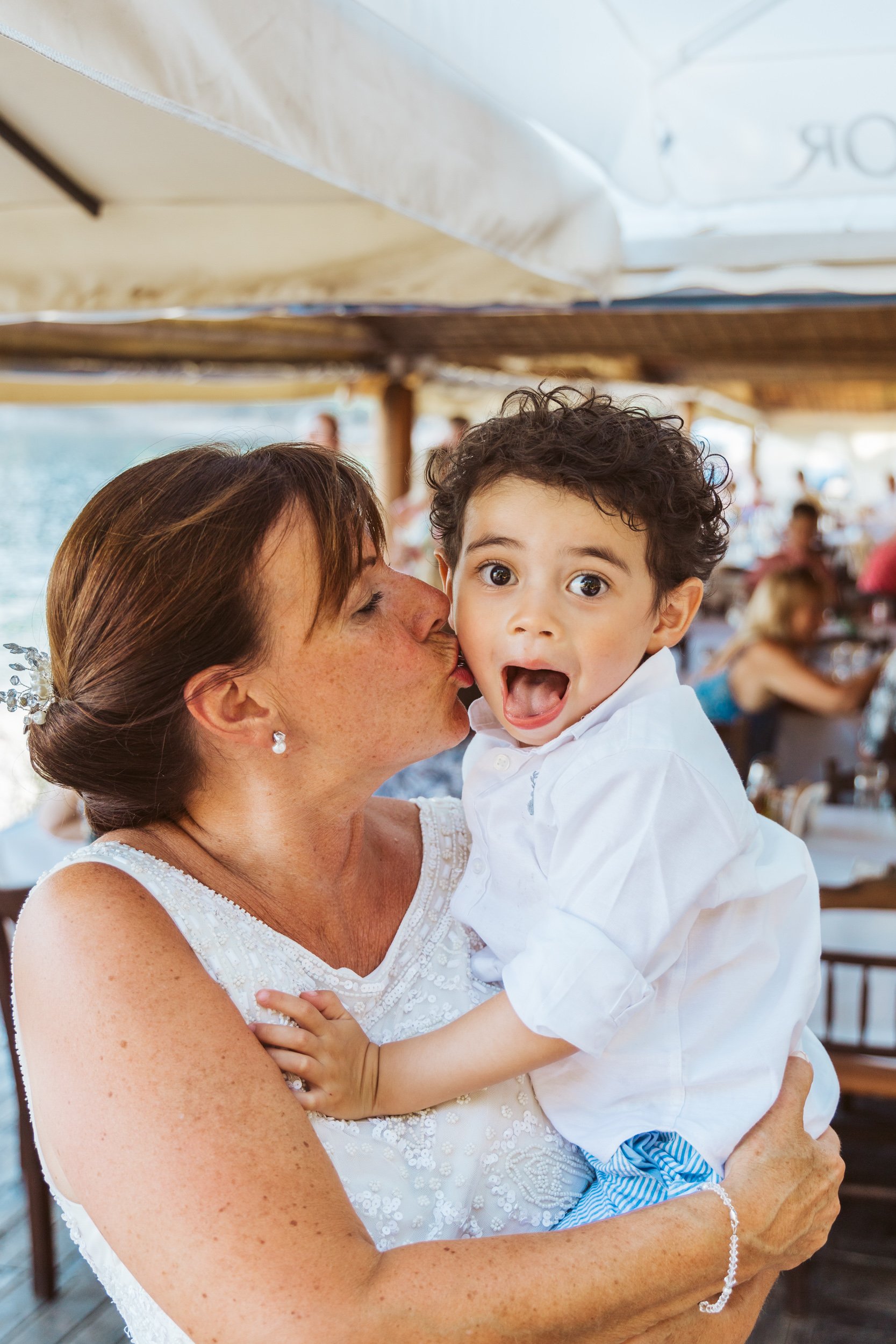 A woman is giving a kiss on the cheek to a young boy with curly hair, who has a surprised expression. They are at an outdoor setting, possibly a restaurant or cafe, with people and tables in the background.