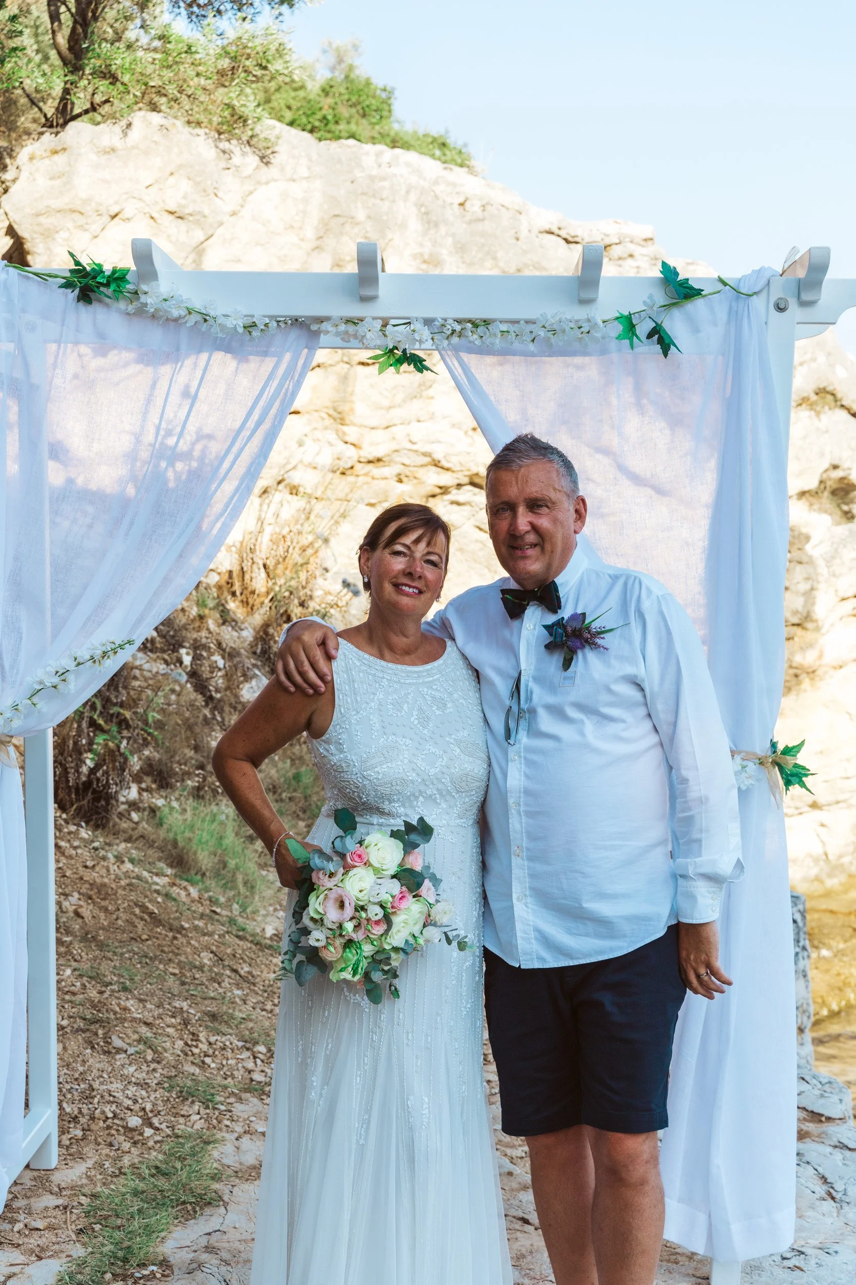 A bride and groom smile and pose under a white wedding arch with sheer curtains and greenery, outdoors with rocky terrain and clear sky in the background.