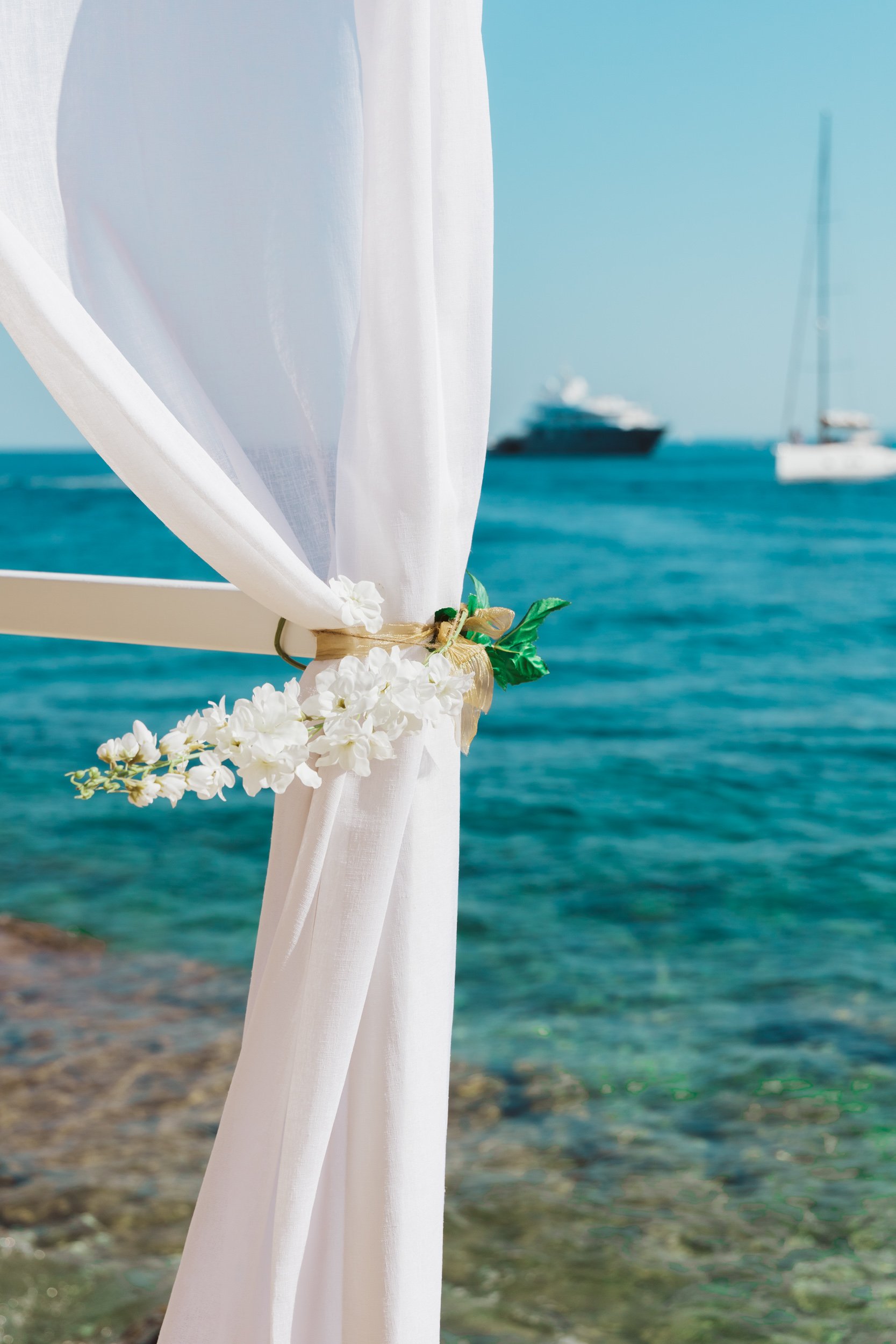 White fabric draped over a wooden structure with decorative white flowers and green leaves, set against a blue ocean background with boats in the distance.