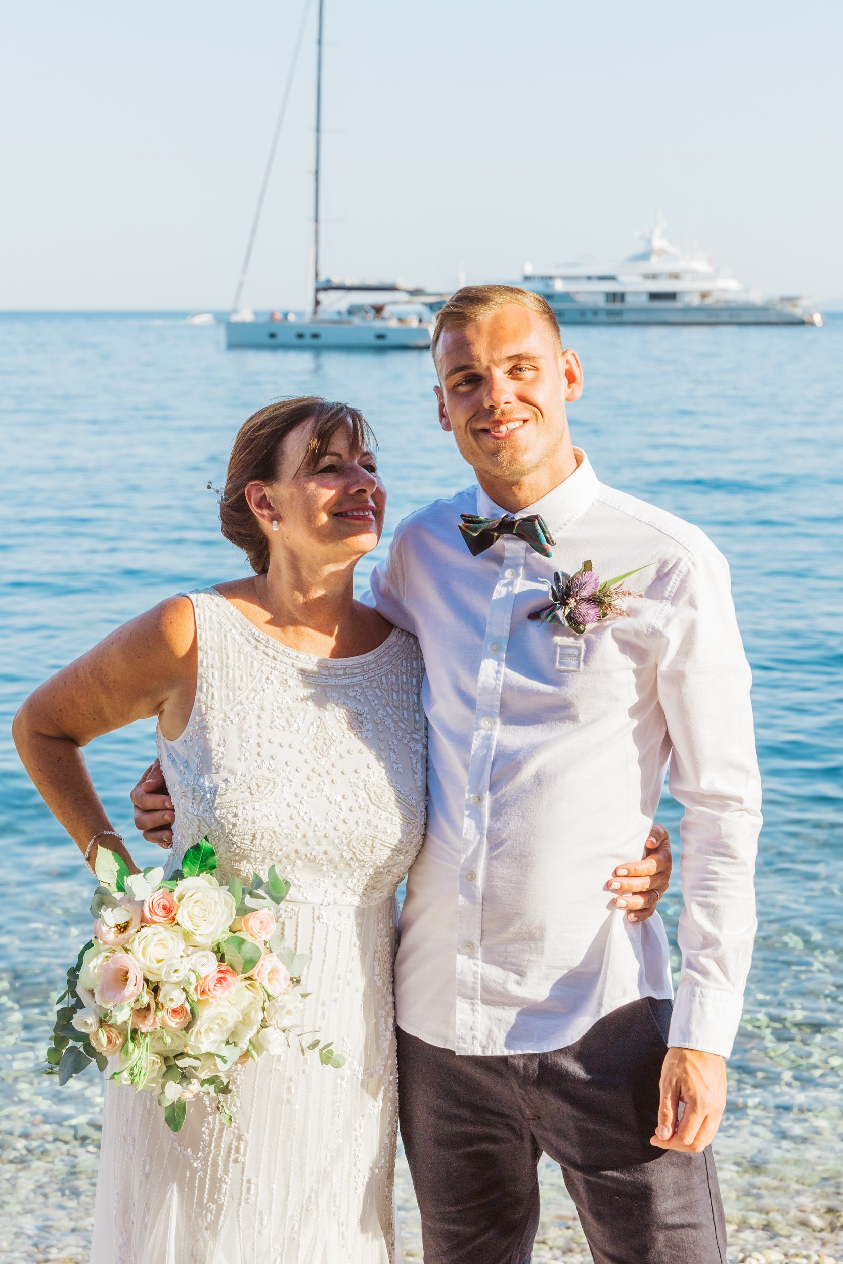 A bride and groom standing on a beach with a large yacht in the background, smiling and embracing each other after their wedding.