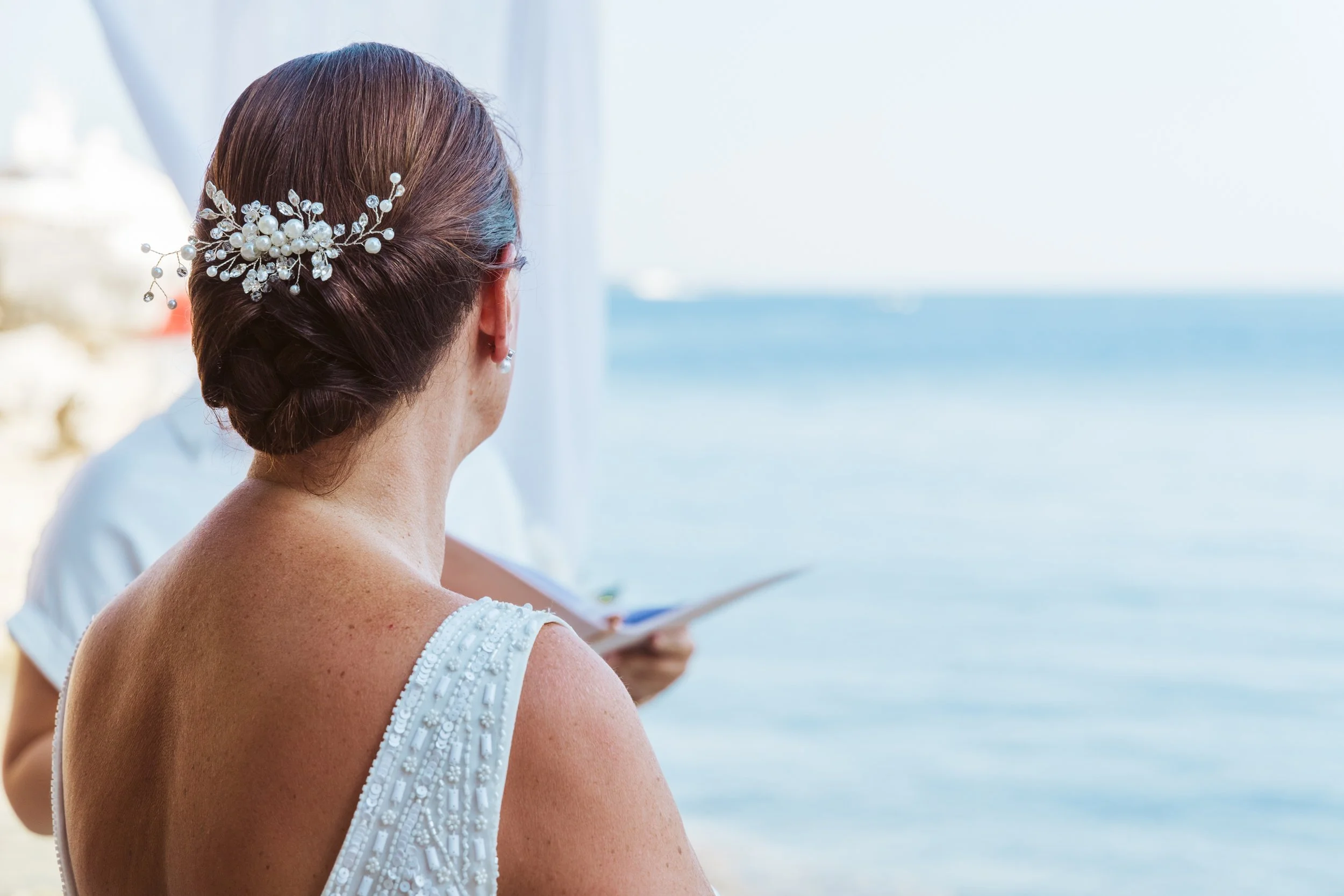 A woman with brown hair in a bun wearing a pearl and flower hairpiece and a sleeveless dress, looks out at the ocean through a window or open space.