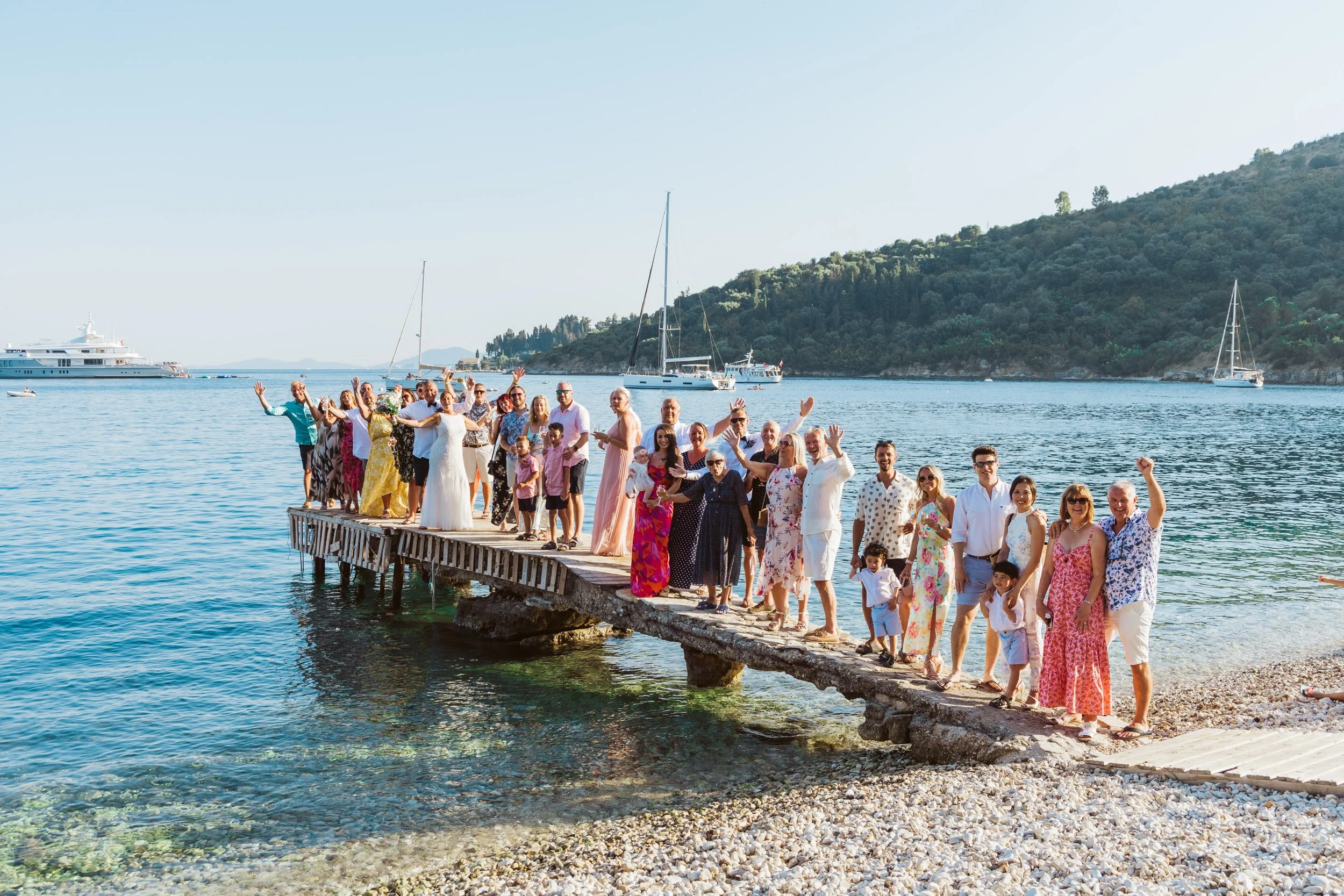 A group of people gathered on a pier by the water, celebrating with their hands raised, with boats and a hill in the background.