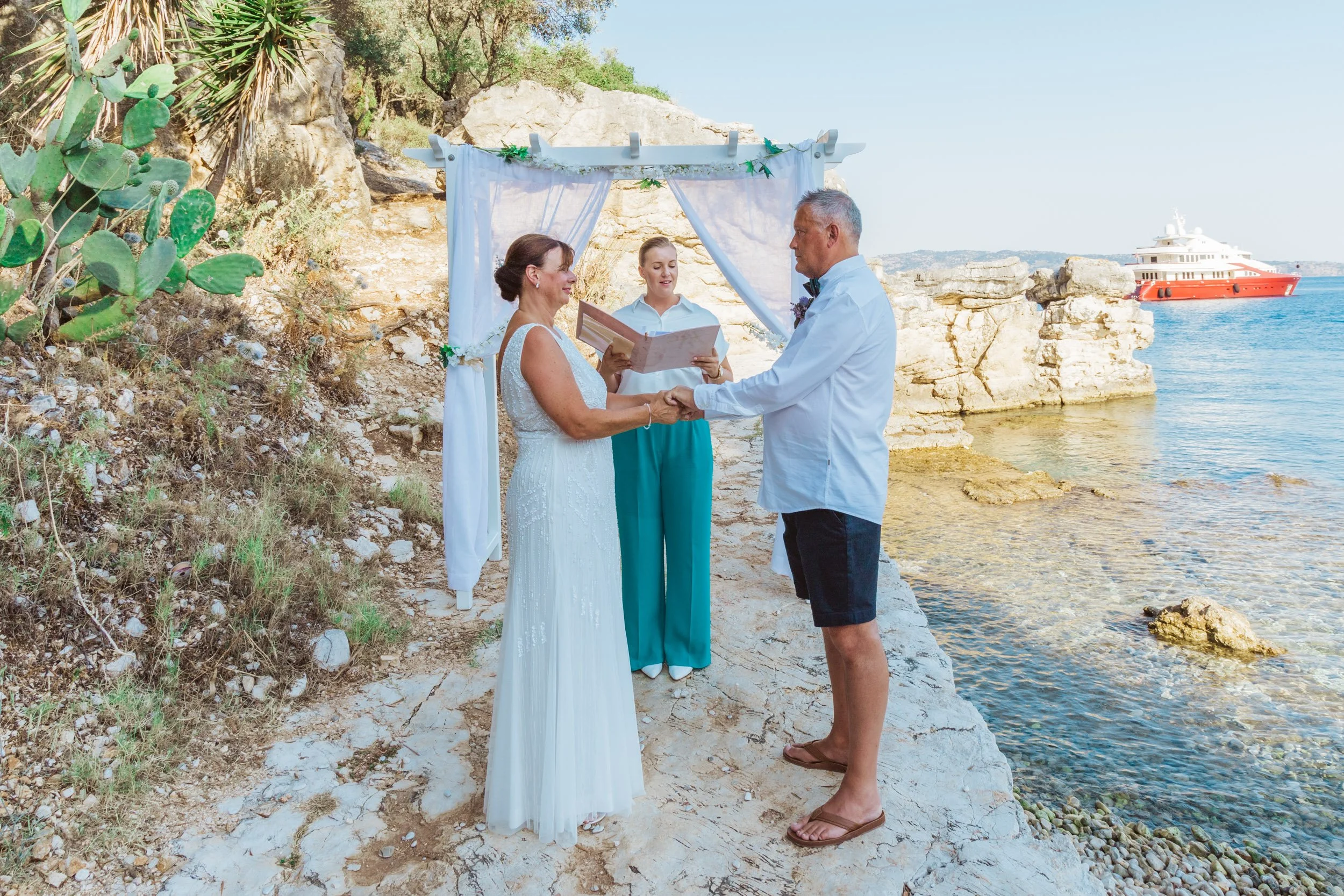 A couple gets married by the water with an officiant, a woman holding a book, under a white canopy with a scenic ocean view and a yacht in the background.