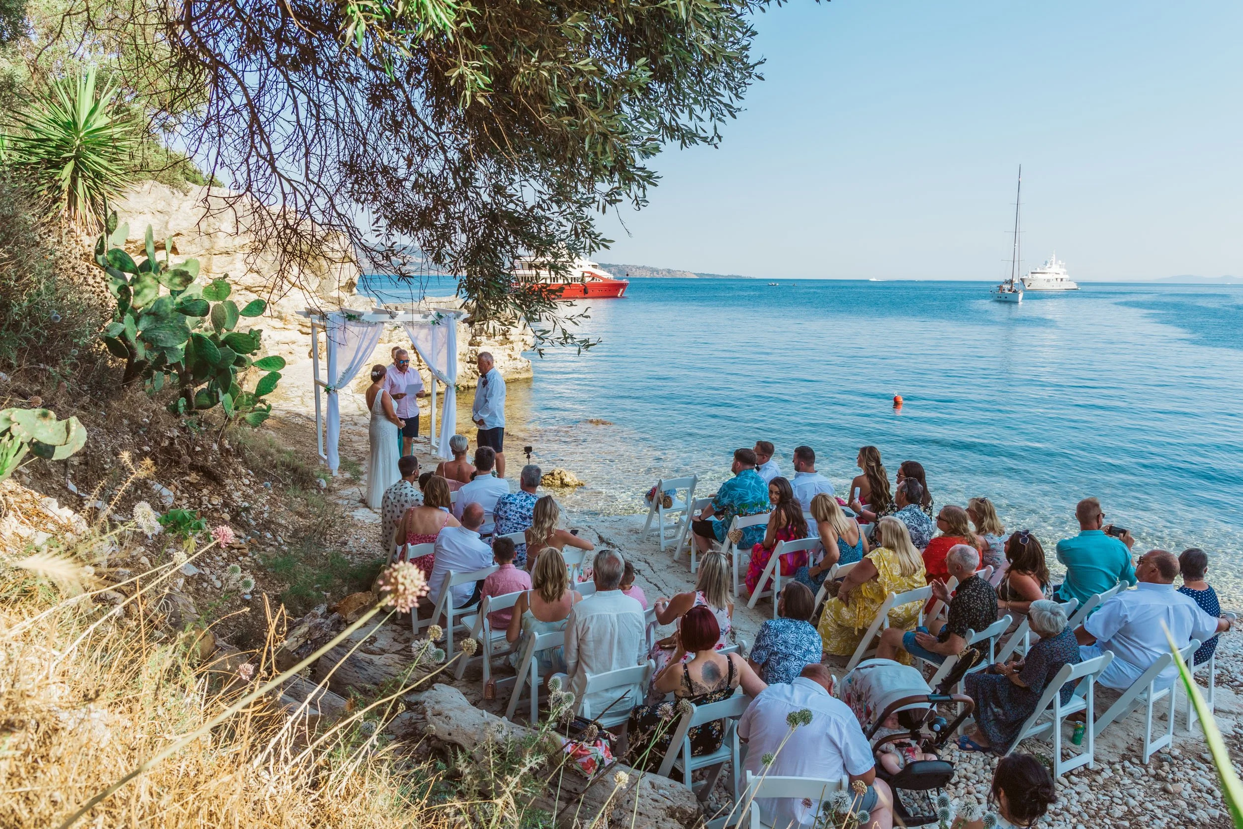A seaside wedding ceremony with guests seated on white chairs on a rocky beach, overlooking blue water with boats, and a wedding officiant and bride and groom standing under a white canopy.