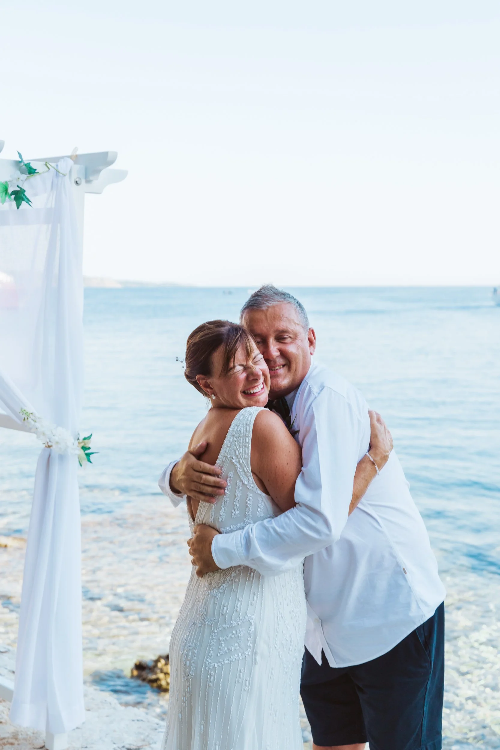 A bride and groom hugging and smiling on a beach during their wedding ceremony with an ocean in the background.
