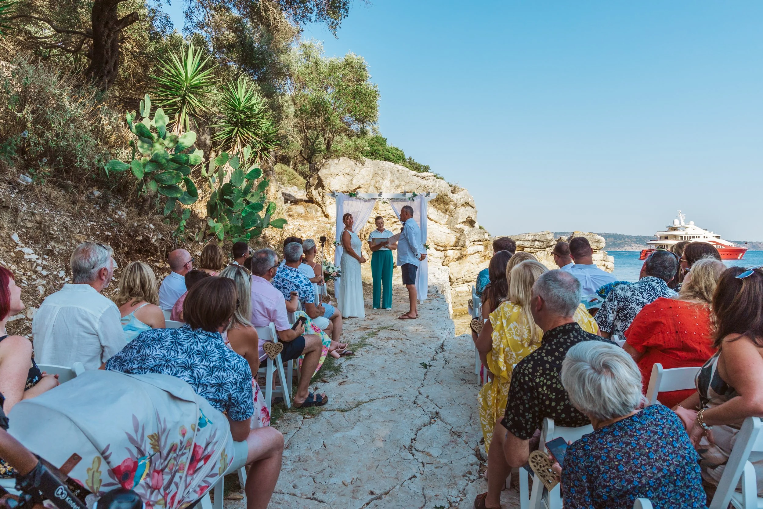 A wedding ceremony taking place outdoors on a rocky beach with a cliff in the background and a boat on the water. The couple is standing under a white canopy, exchanging vows with an officiant while guests seated in white chairs watch. There are cact