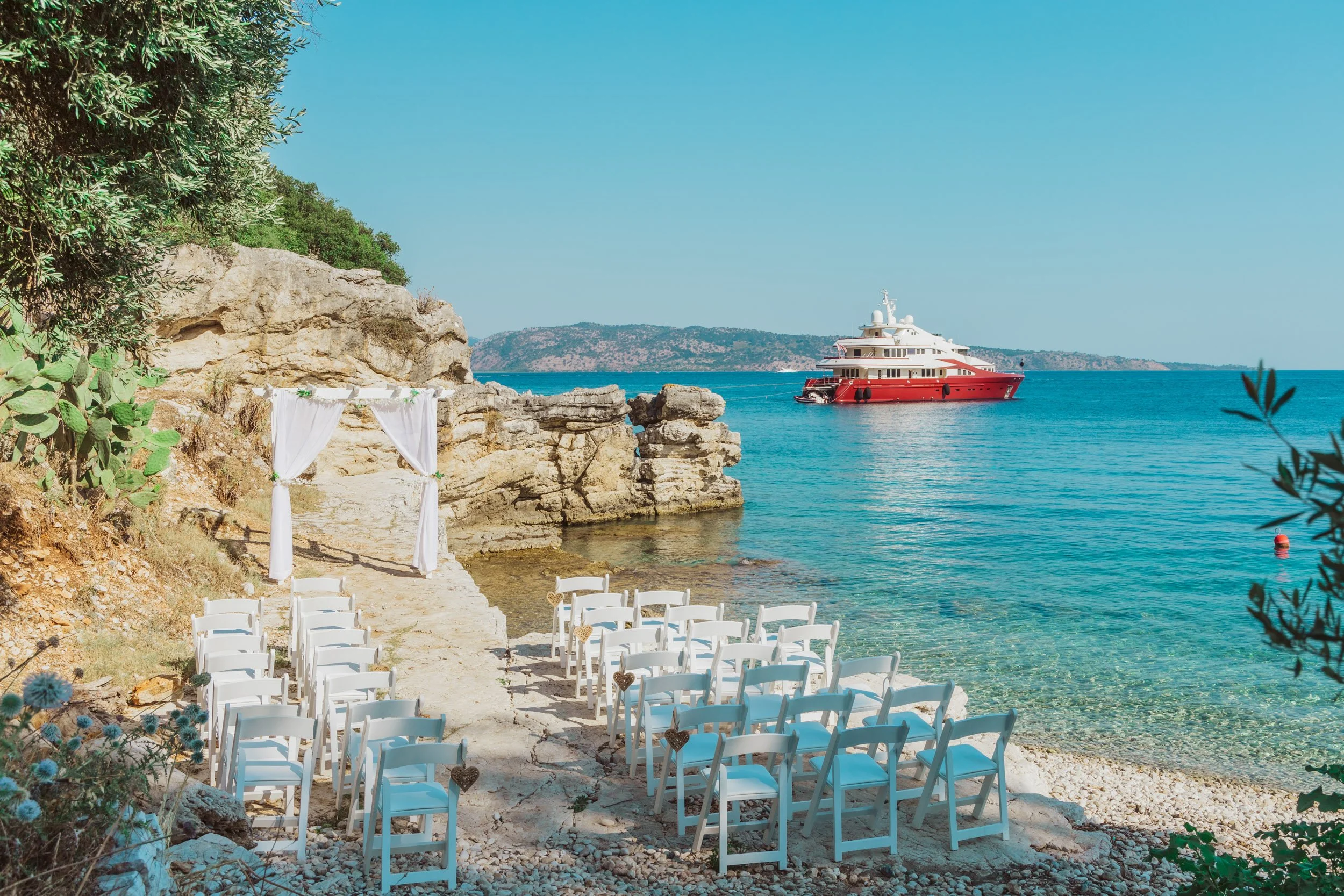 A seaside wedding setup with white chairs arranged in front of a white draped arch on a rocky beach, overlooking clear blue water and a red yacht, with distant hills under a bright blue sky.