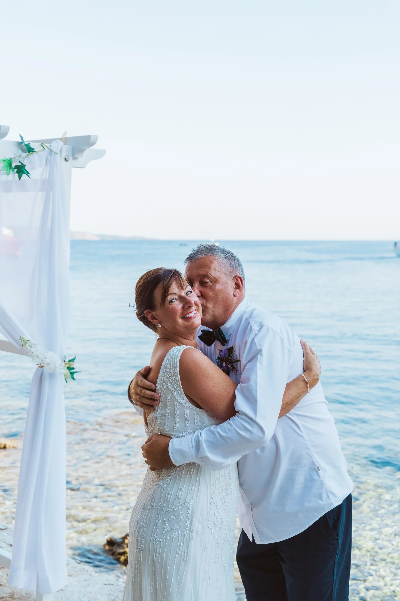 An older couple in wedding attire hugging by the ocean, with a wedding arch decorated with white fabric and greenery in the background.