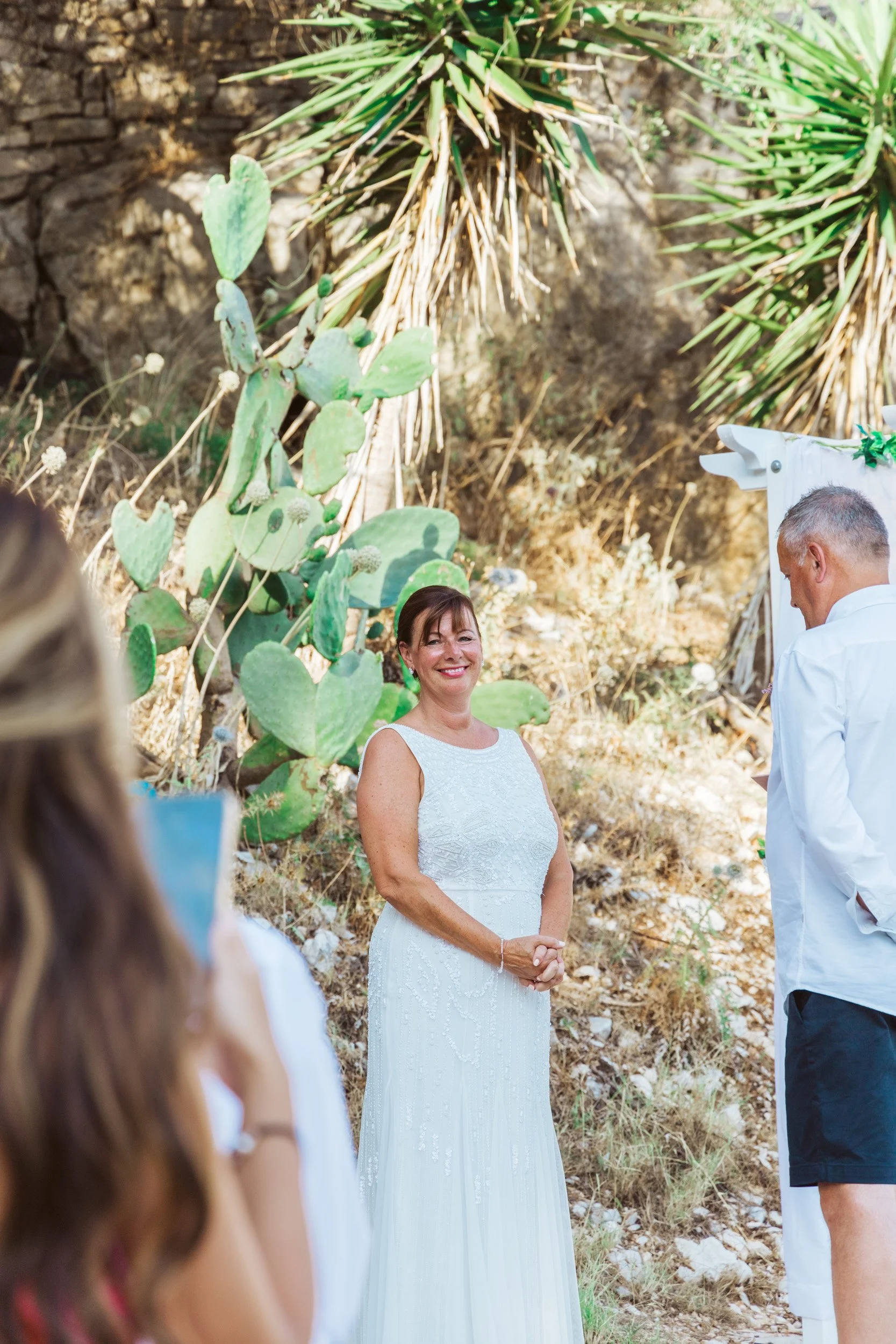 A woman in a white dress standing outdoors near cacti and desert plants, smiling during what appears to be a wedding or celebration.