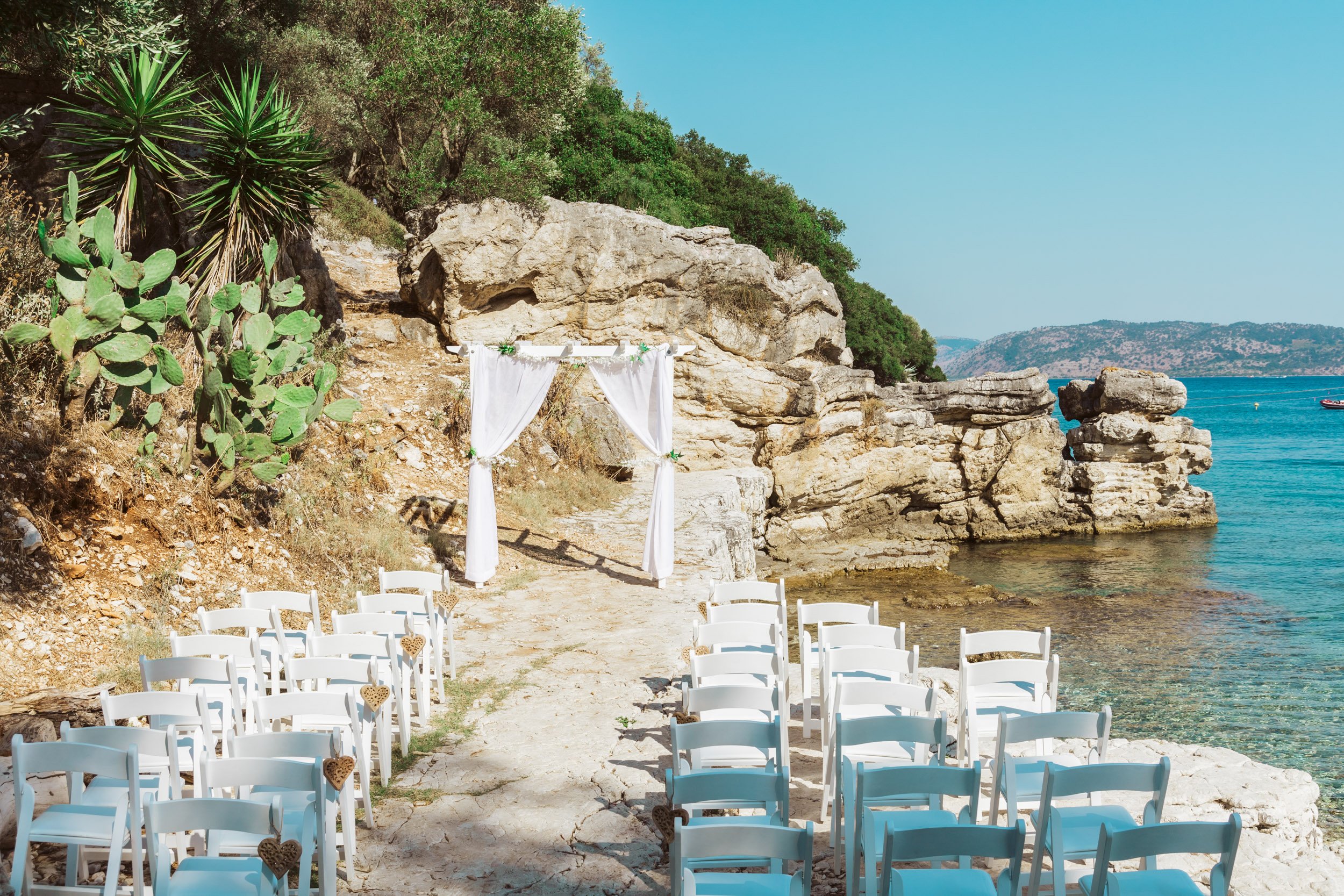 A seaside wedding setup with white chairs decorated with heart-shaped ornaments, facing an arch draped with white fabric, on a sandy beach with rock formations and trees, overlooking the ocean with boats and distant hills.