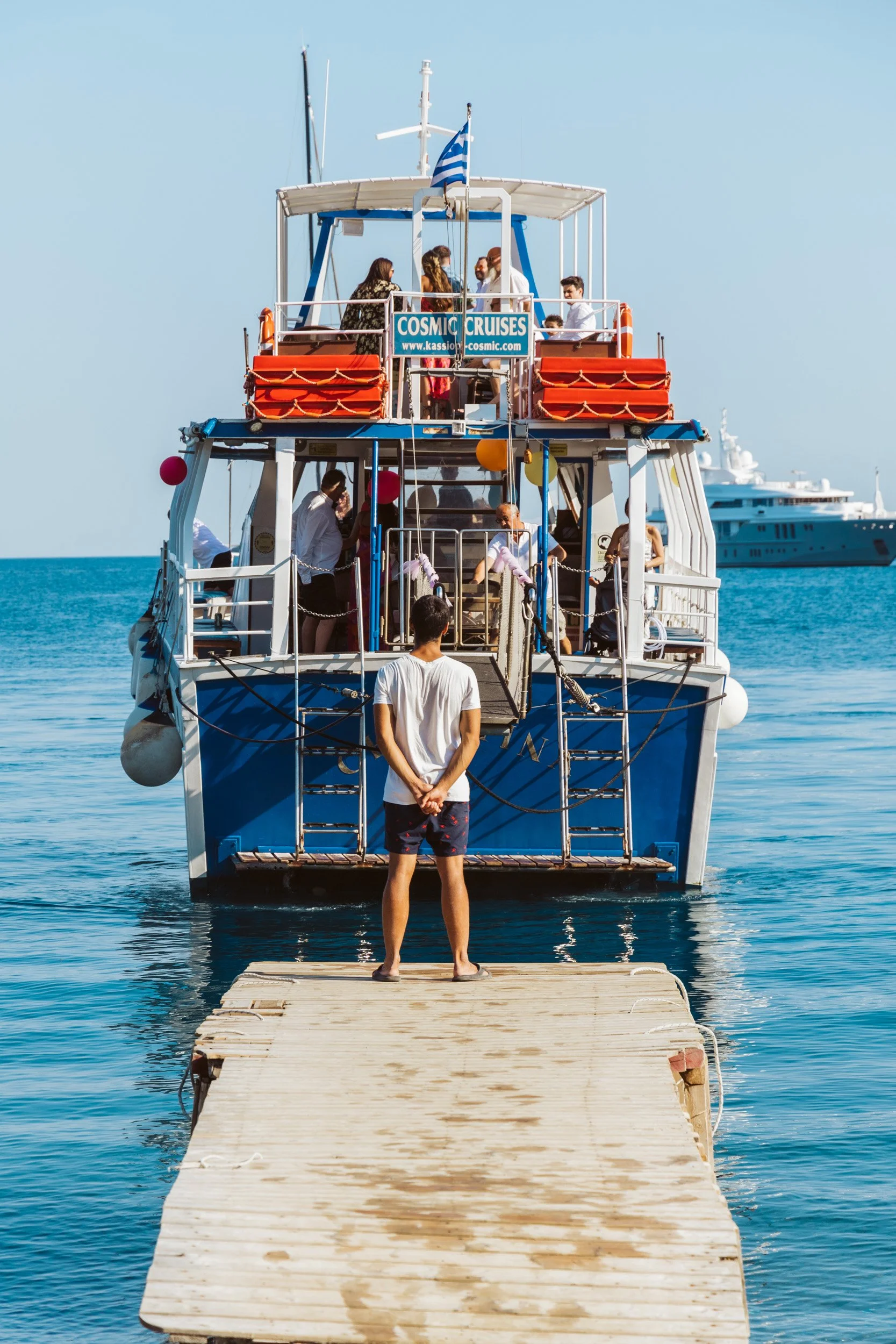 A man stands on a wooden dock looking at a large blue and white cruise boat named Cosmic Cruises, docked in clear blue water, with other boats in the background, on a sunny day.