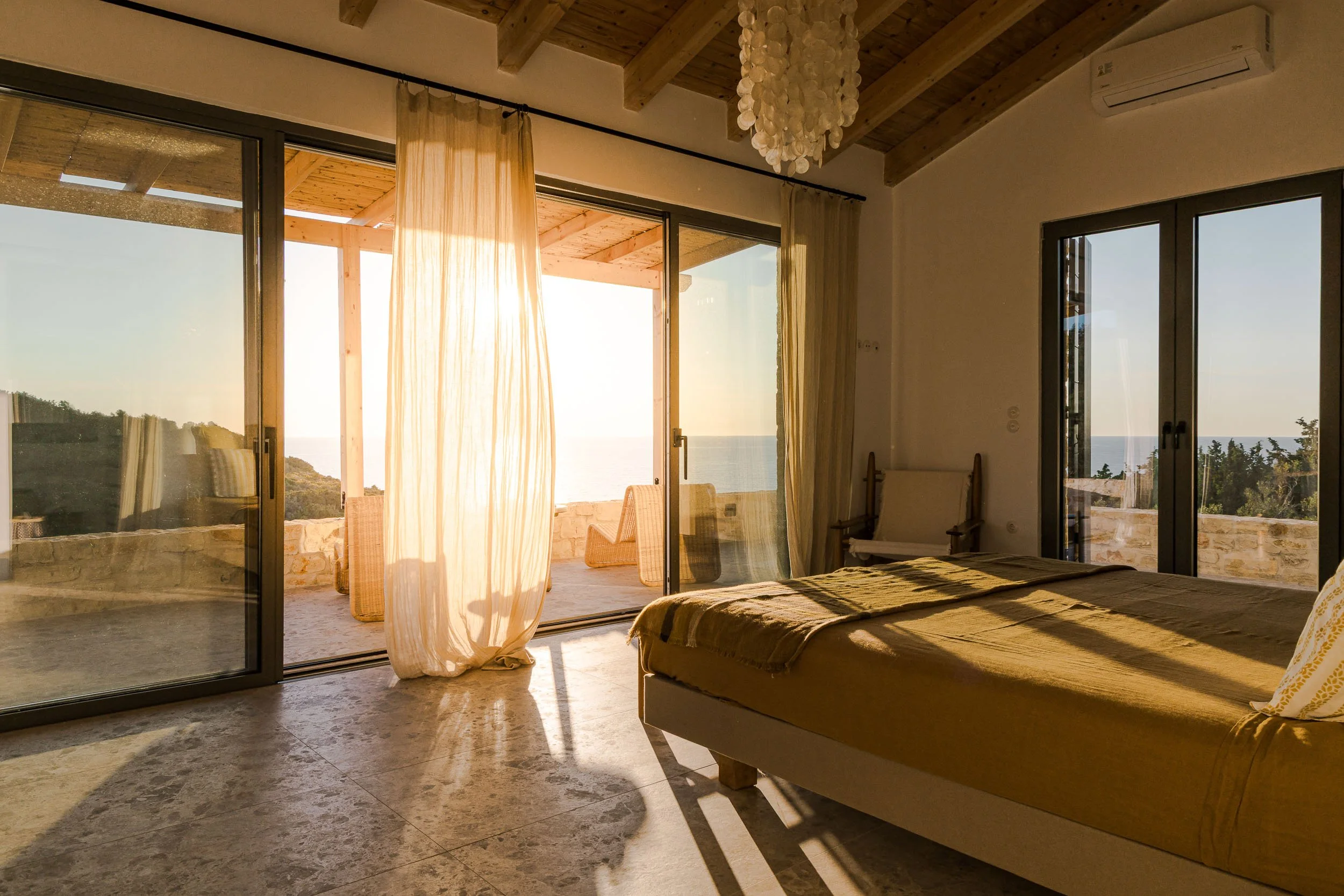 A bedroom with beige bedding and large glass doors that open to a balcony with an ocean view, sunlight streaming through sheer curtains.