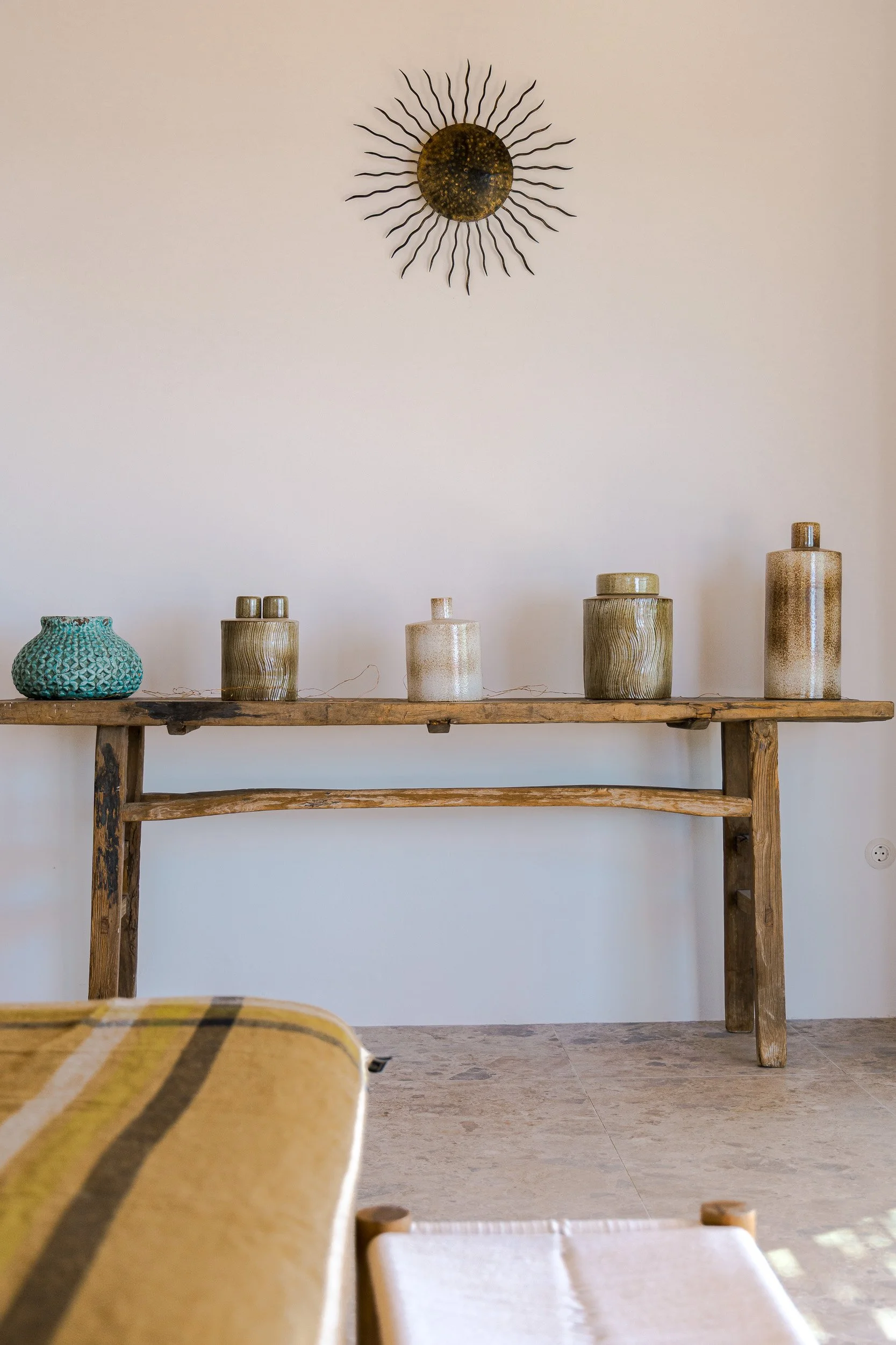 Wooden console table with five ceramic vases of varying heights and colors, a decorative orb, against a plain white wall with sunburst wall art.