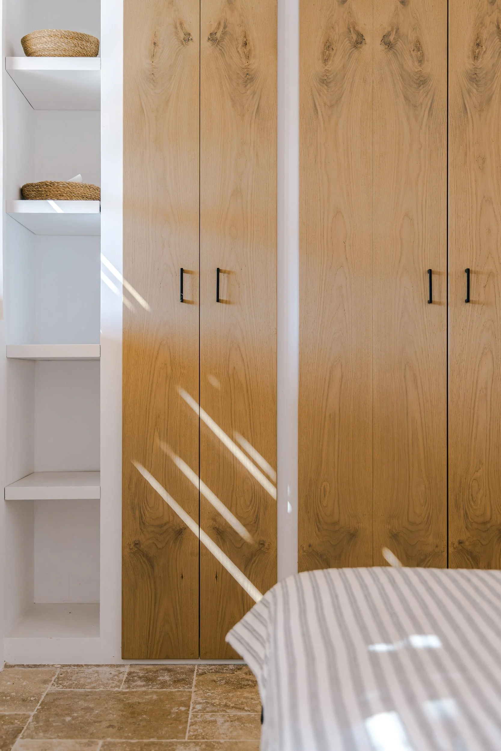 Wooden closet doors with black handles, white open shelves on the left with baskets, sunlight casting diagonal shadows on the closet, a striped fabric covering part of a bed or chair in the foreground, beige tiled floor.