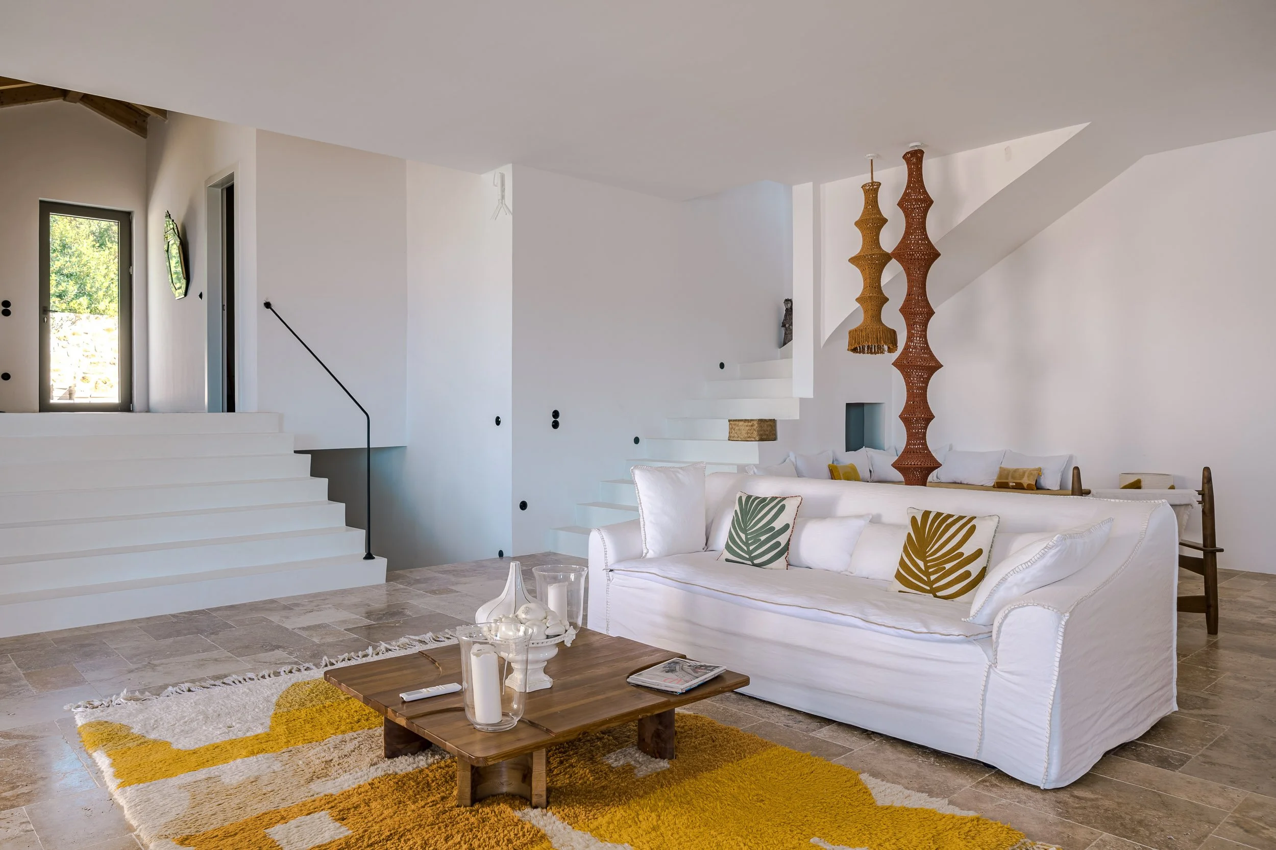 Living room with white sofa, yellow and green leaf pillows, wooden coffee table with candles, yellow rug, stairs, and decorative hanging lamps.