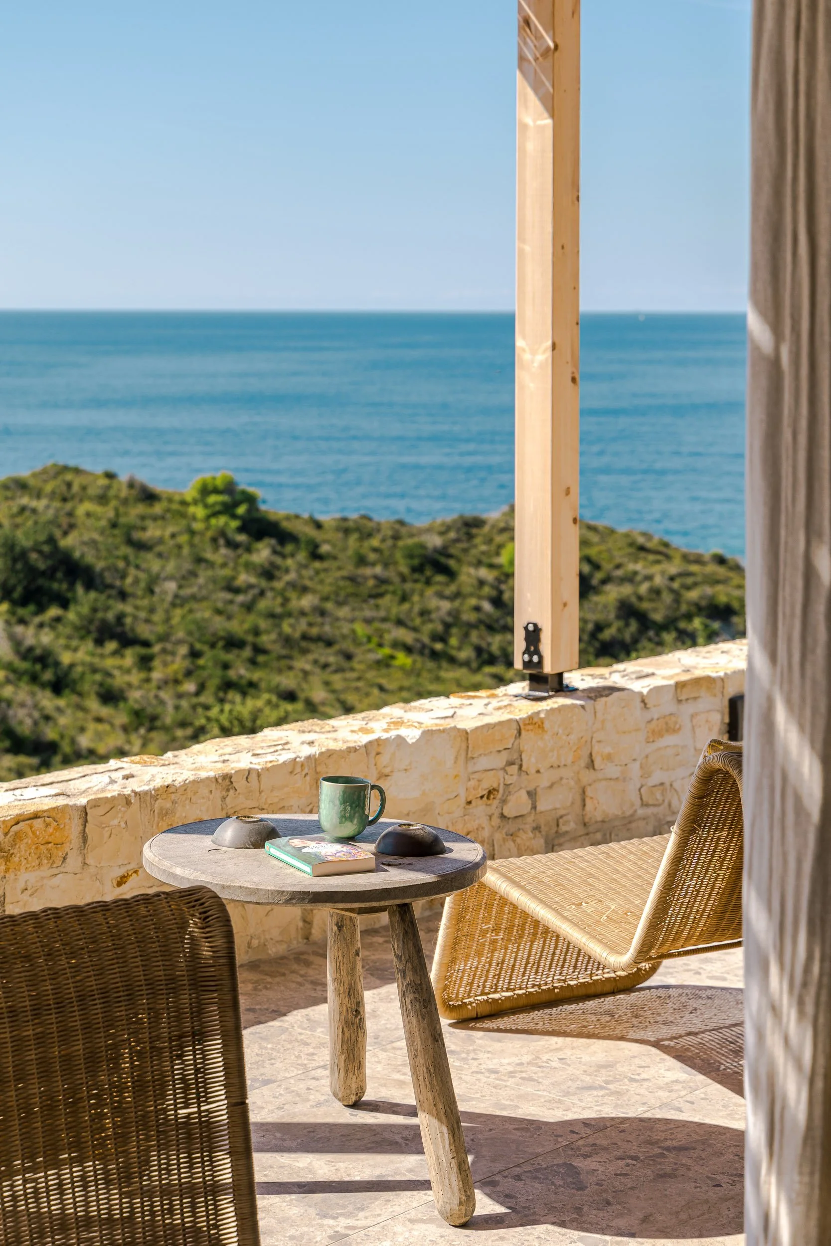 A balcony overlooking the ocean with a small round table, a book, a green mug, and two black bowls. There is a wicker chair and a lounge chair on the balcony.