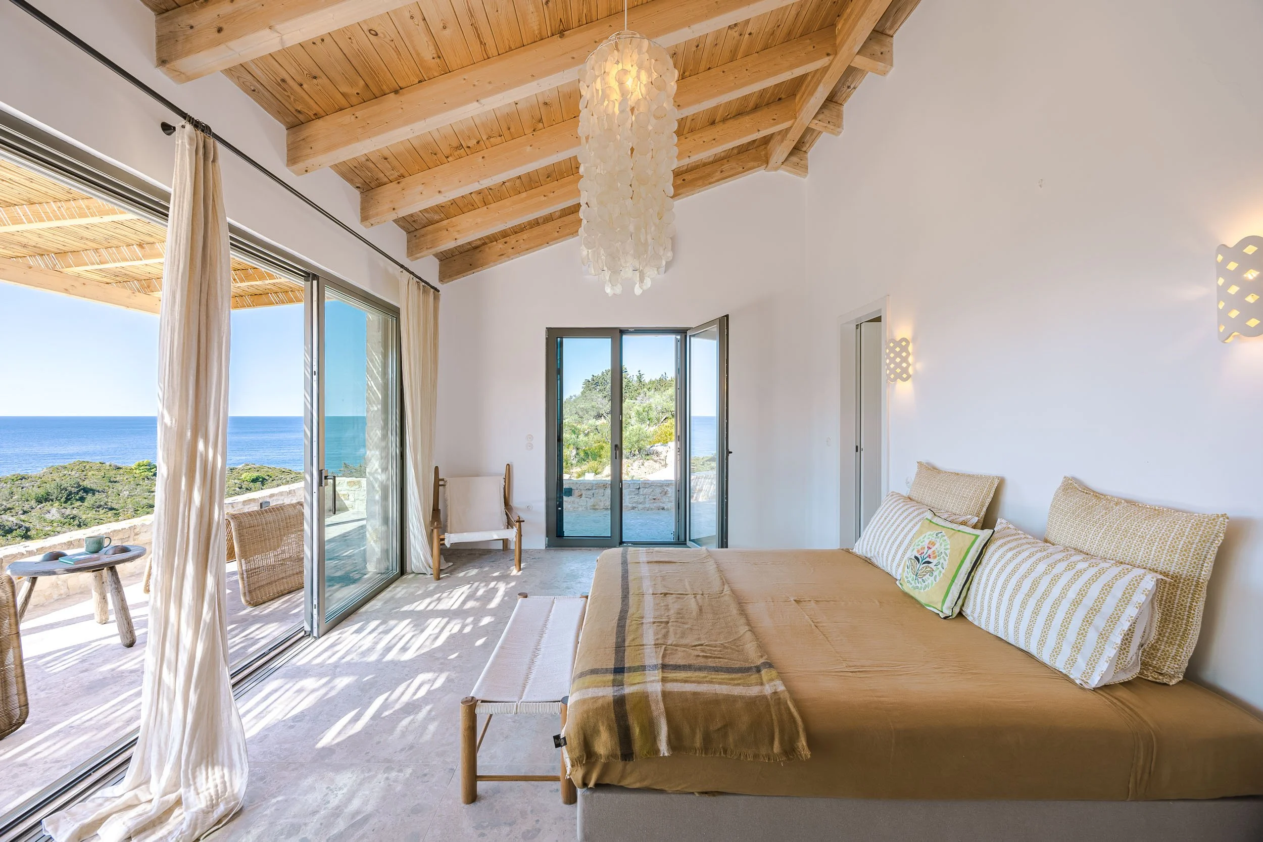 Modern bedroom with large sliding glass doors leading to an outdoor patio with ocean view, wooden ceiling, bed with beige bedding and patterned pillows, and cozy chair near the door.