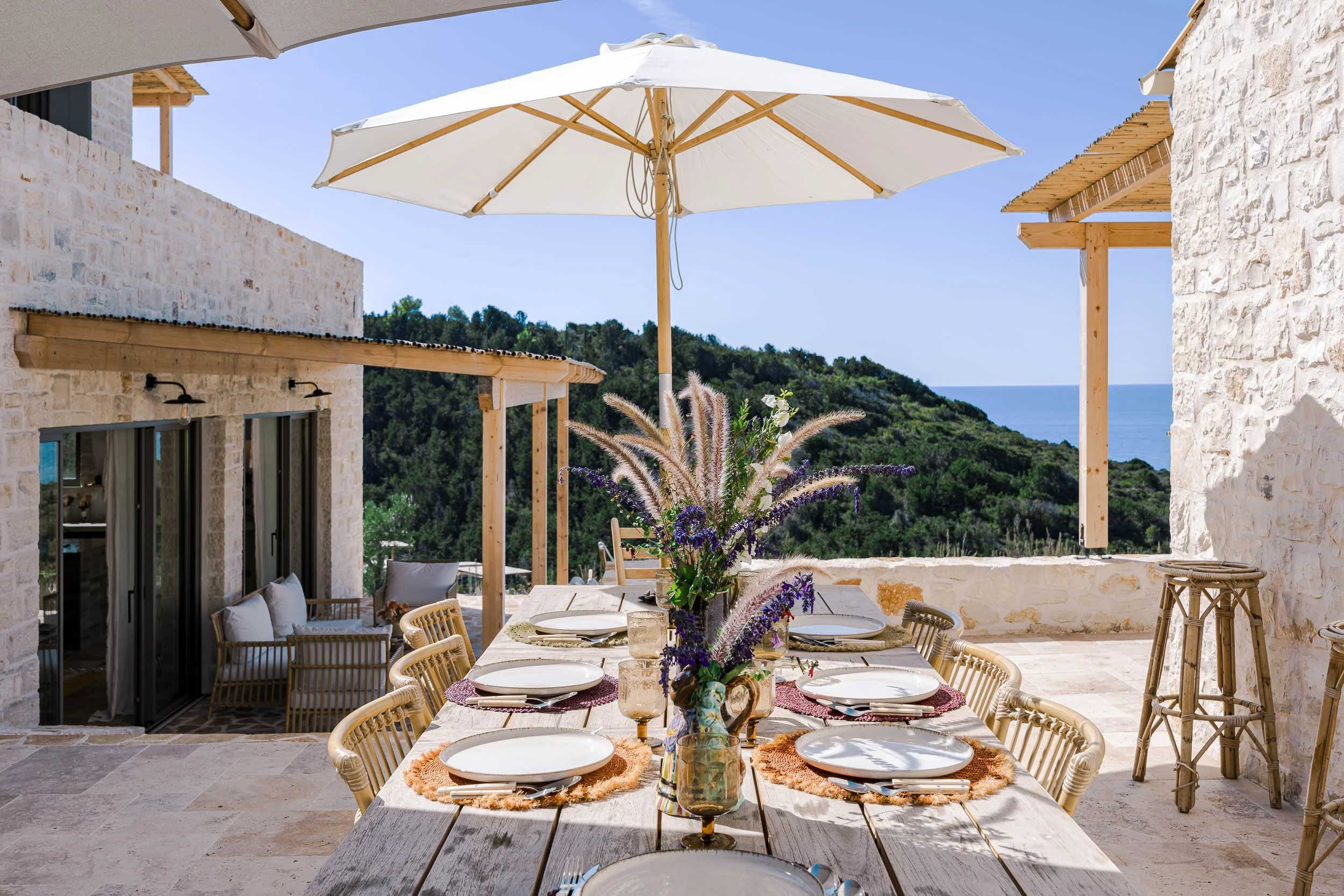 Outdoor dining table set for six with white plates, glasses, and a floral centerpiece under a white patio umbrella, overlooking a hillside and ocean.