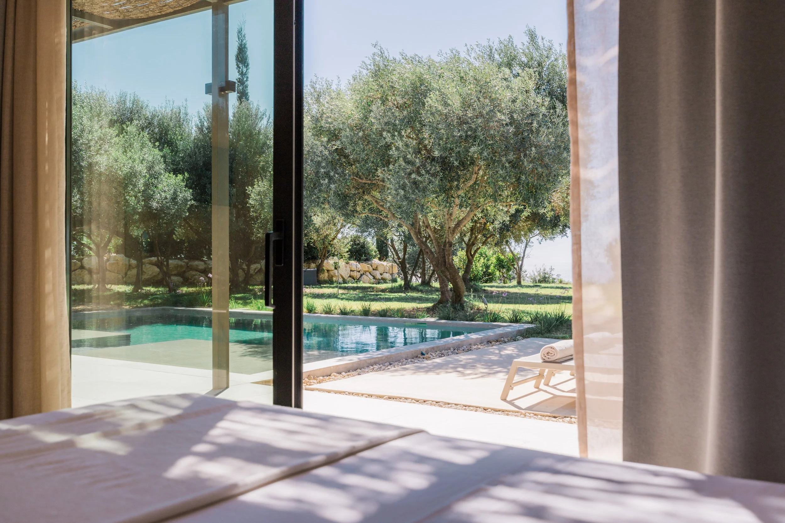 View through sliding glass door of backyard with swimming pool, trees, and lawn, with a white lounge chair and rolled towel outside.