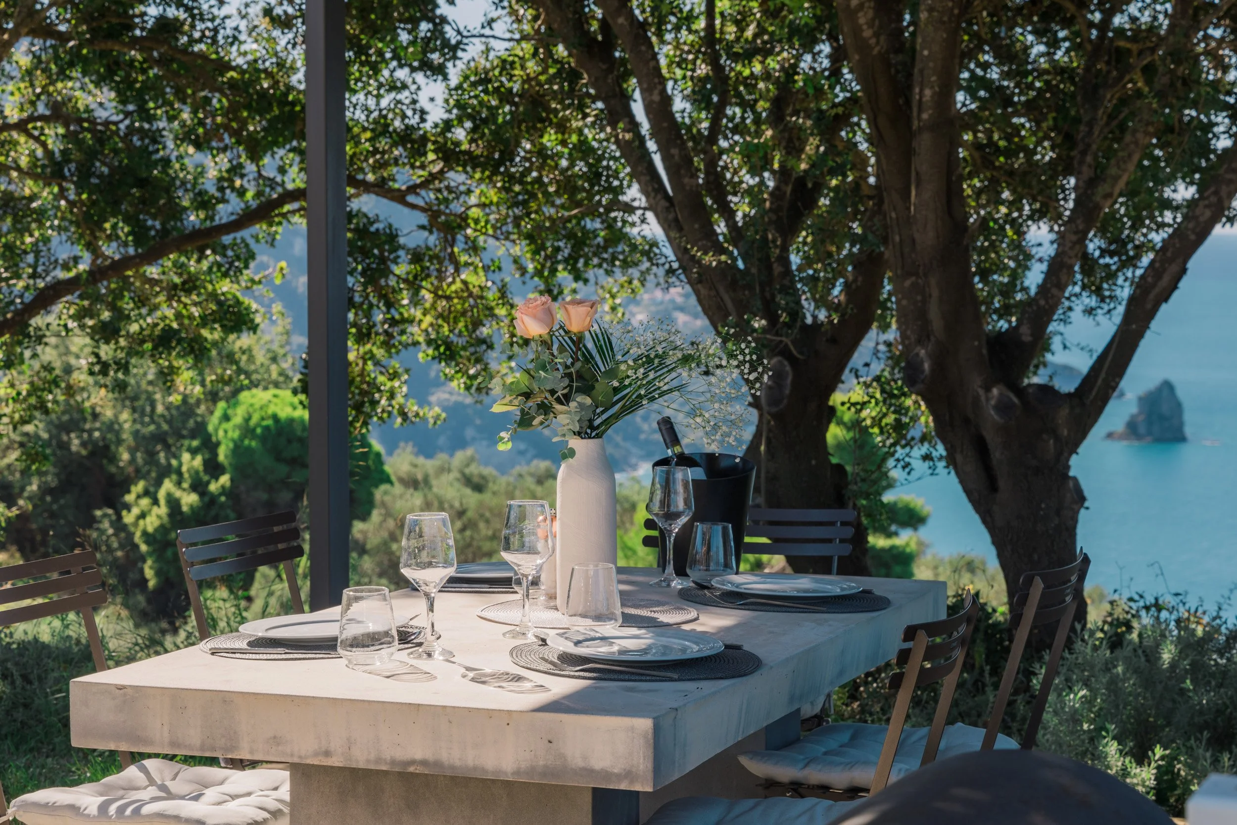 Outdoor dining table set with glasses, plates, a vase of pink roses, and a bottle of wine in an outdoor setting with lush green trees and ocean view in the background.