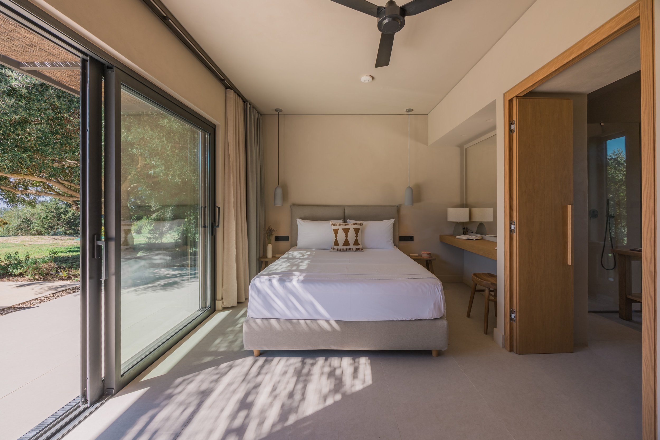 Modern hotel room with a bed, side tables, pendant lights, a desk, a wooden stool, and sliding glass doors opening to a garden with trees and grass